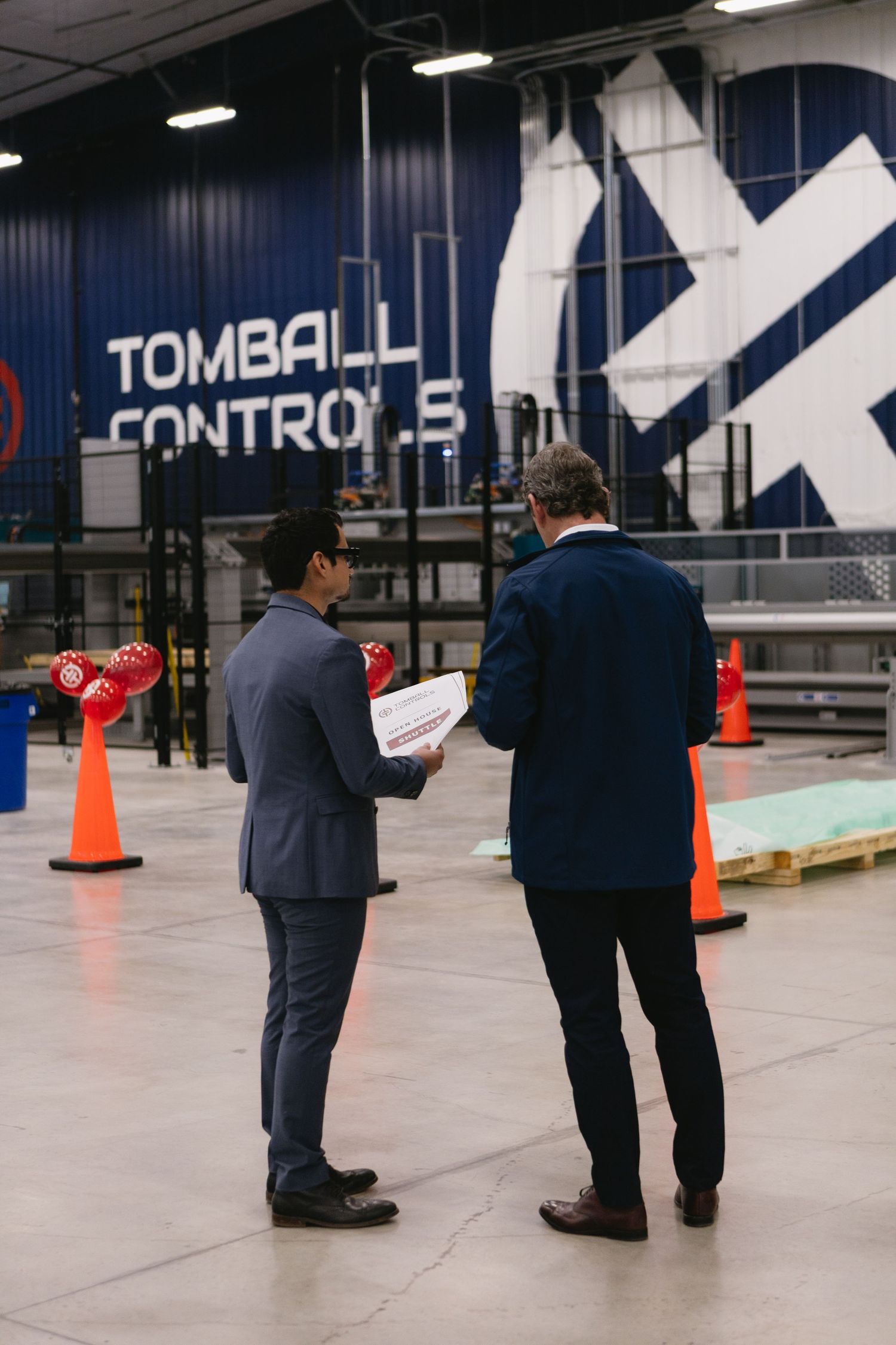 Two people in business attire stand together examining documents in a large industrial warehouse space.