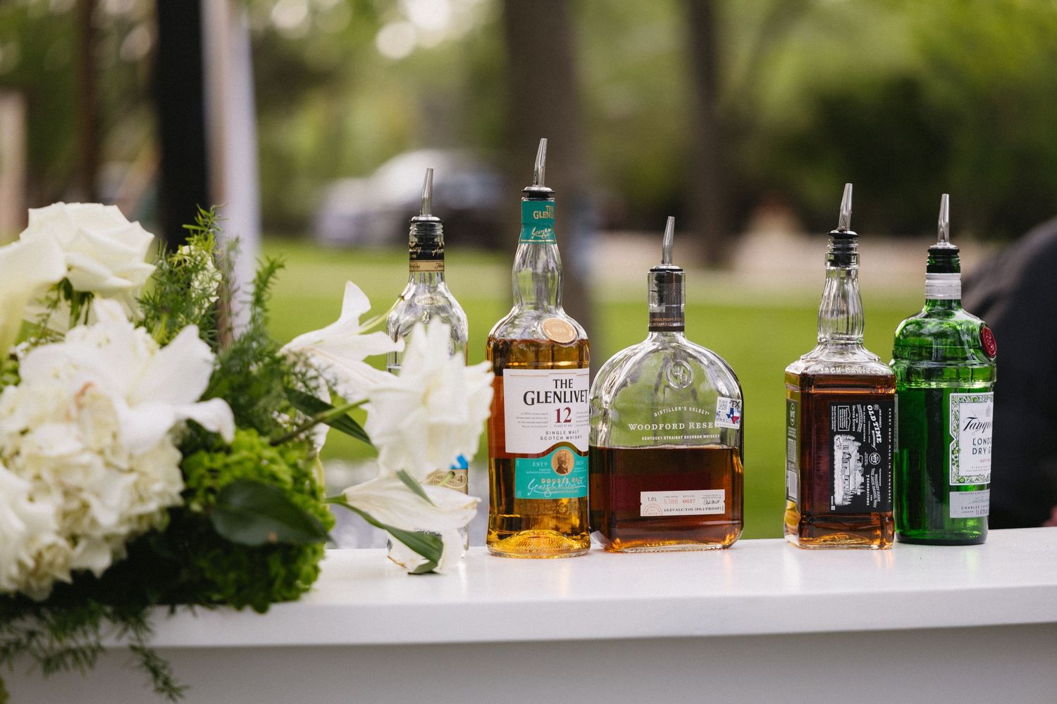 Assorted liquor bottles including whiskey and gin displayed on a white bar table with white floral arrangement at outdoor wedding.