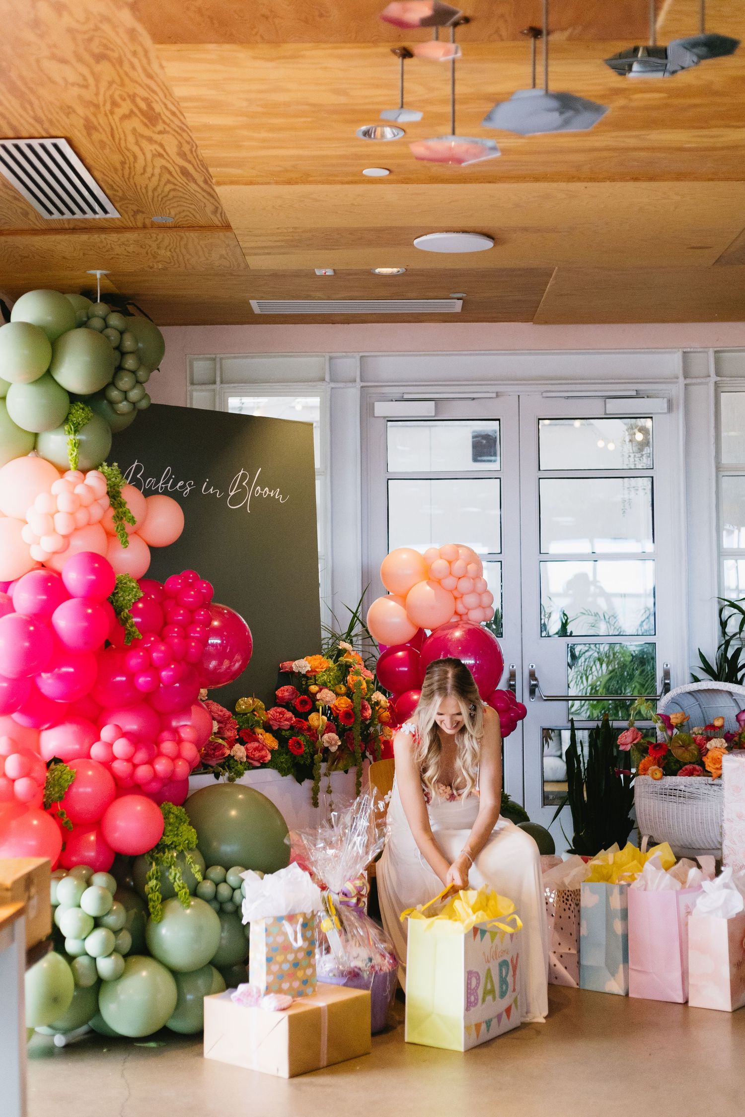 Pregnant woman sits surrounded by colorful balloon garland and gifts at a vibrant baby shower celebration.