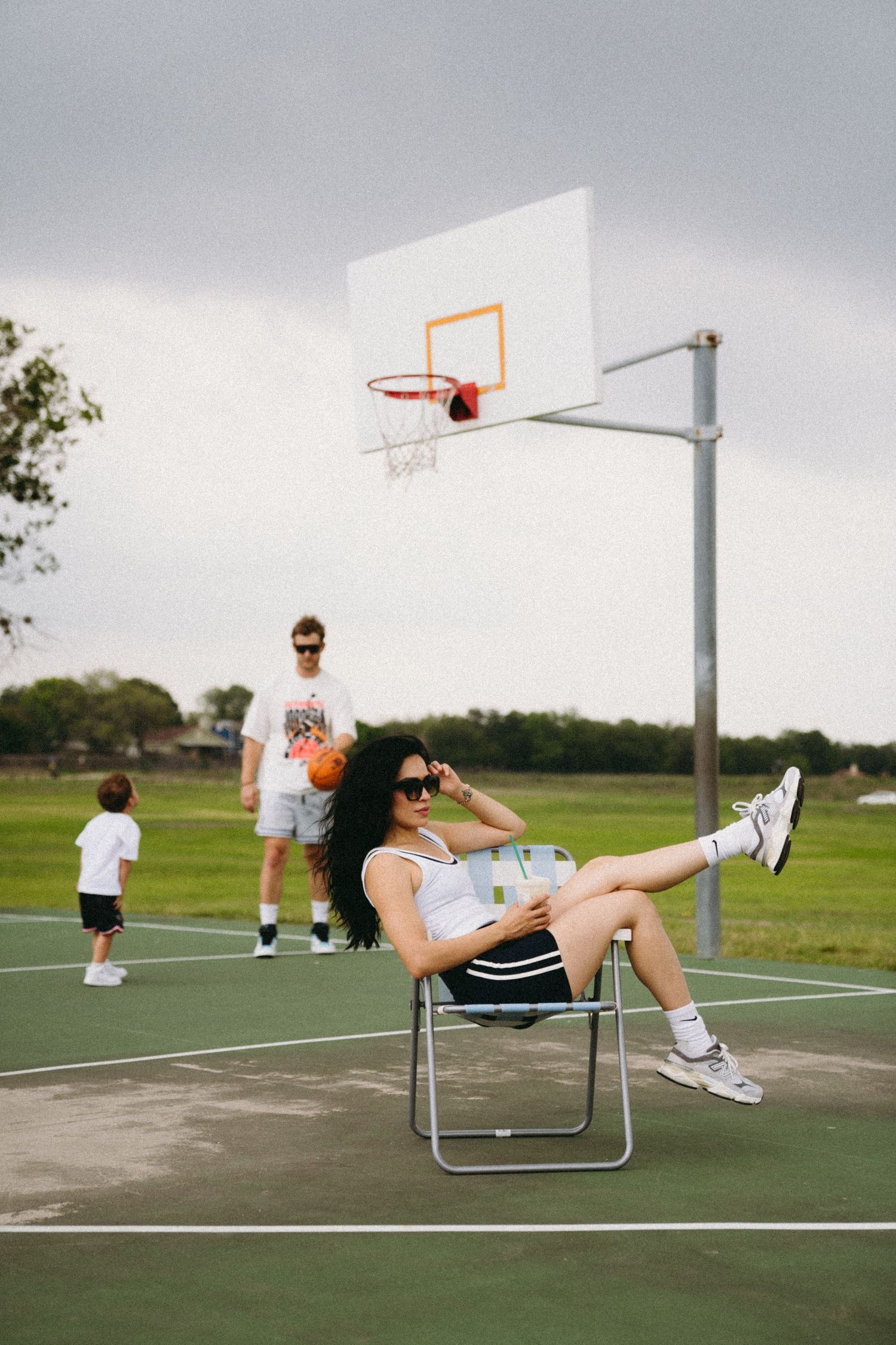 Woman relaxing in a chair on a basketball court wearing sunglasses, while a man and child play in the background.