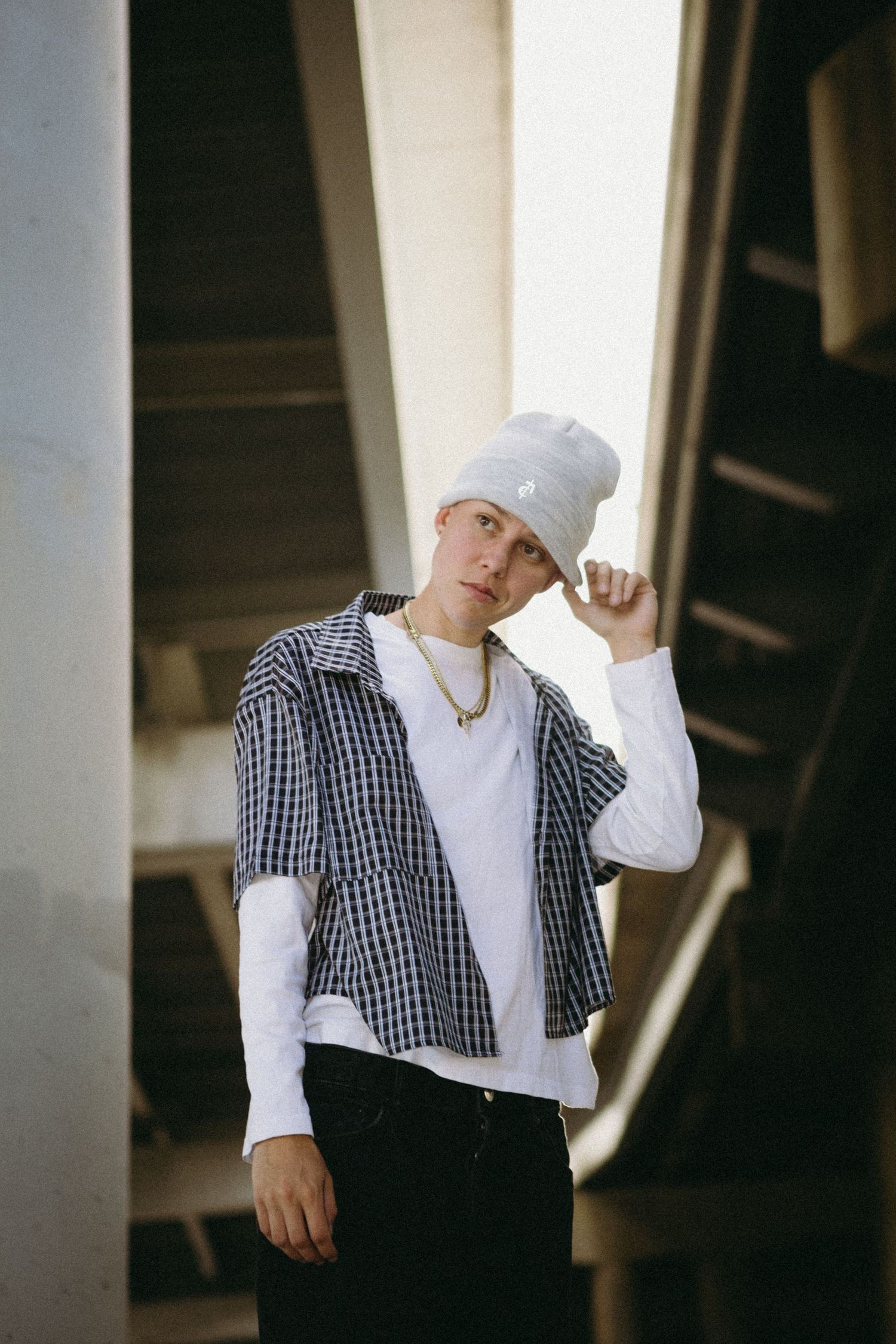 Young man in plaid shirt, white tee, black jeans, and gray beanie poses under urban structure.
