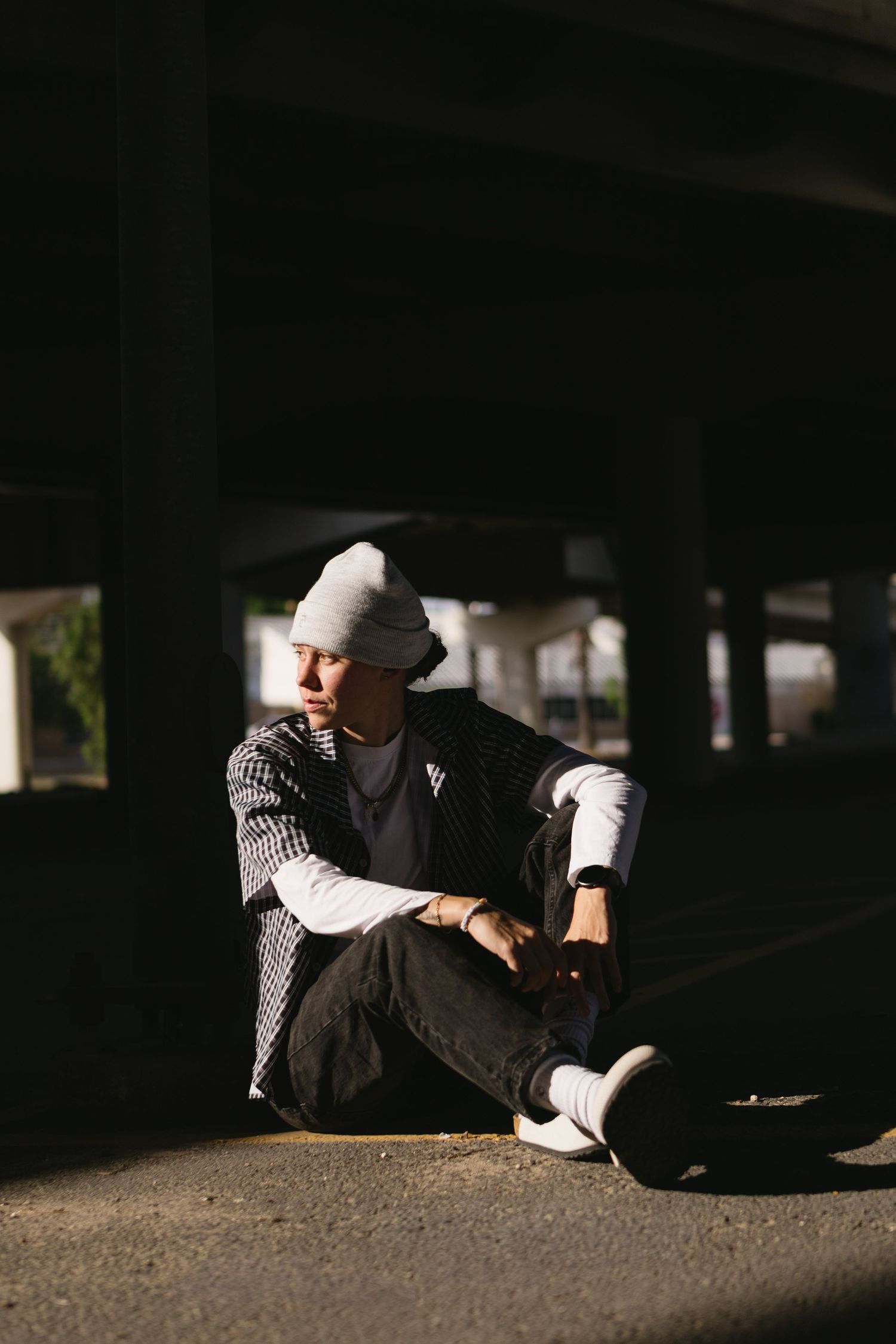 Stylish young man in gray beanie and plaid jacket sitting on ground in dramatic shadow under urban overpass.