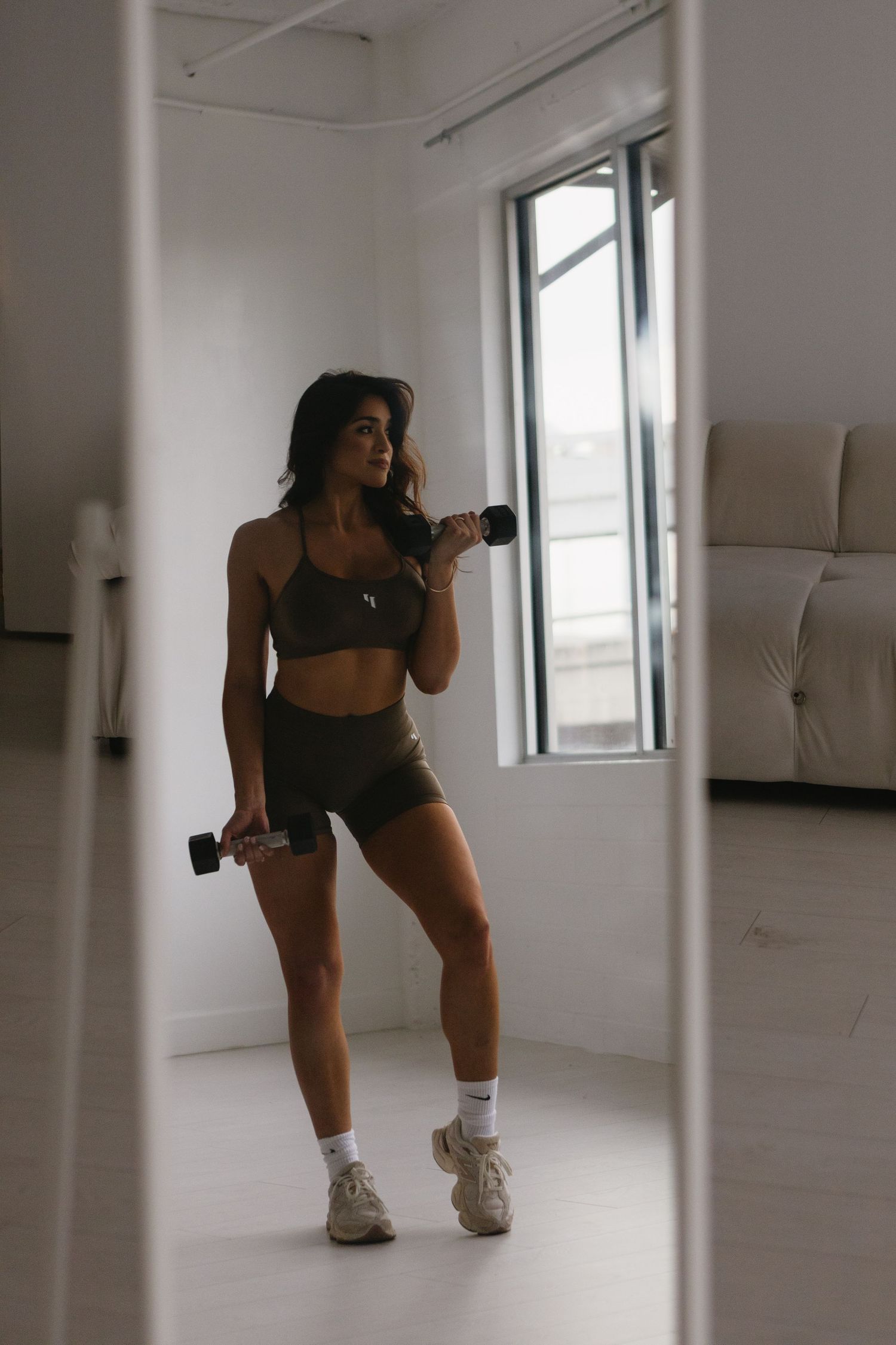 Fit woman in olive green sports bra and shorts doing dumbbell curls in a bright white home gym.