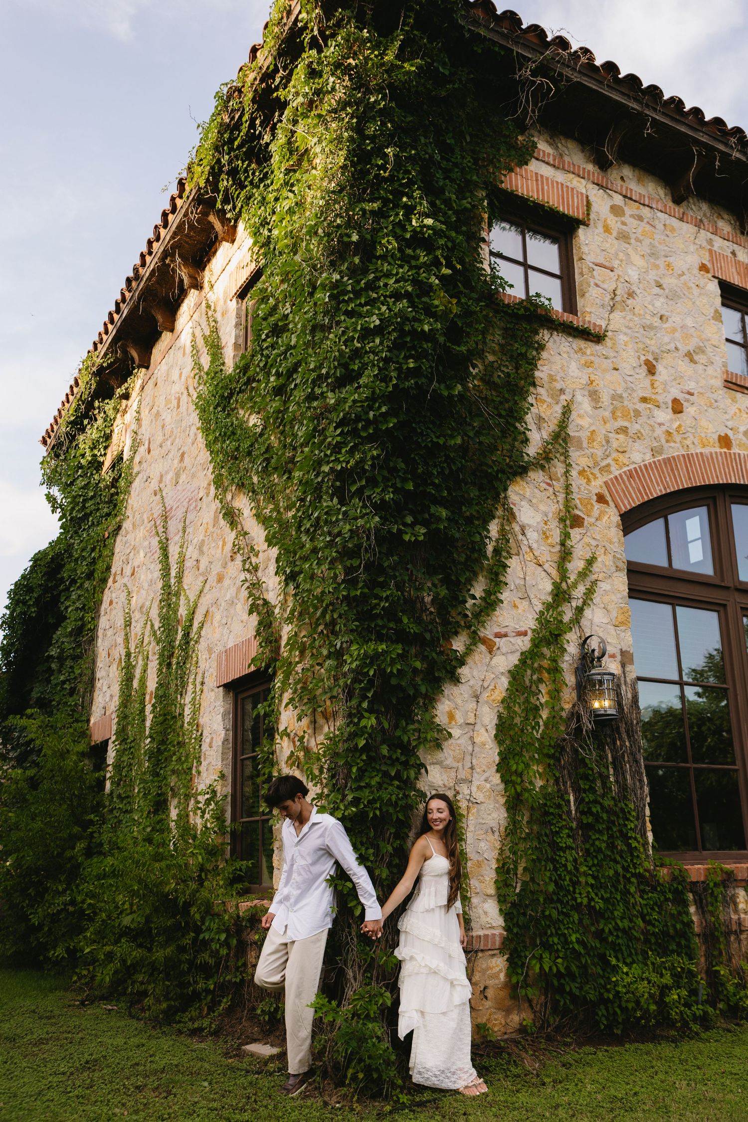 Bride and groom holding hands outside an ivy-covered stone villa, wearing white wedding attire.