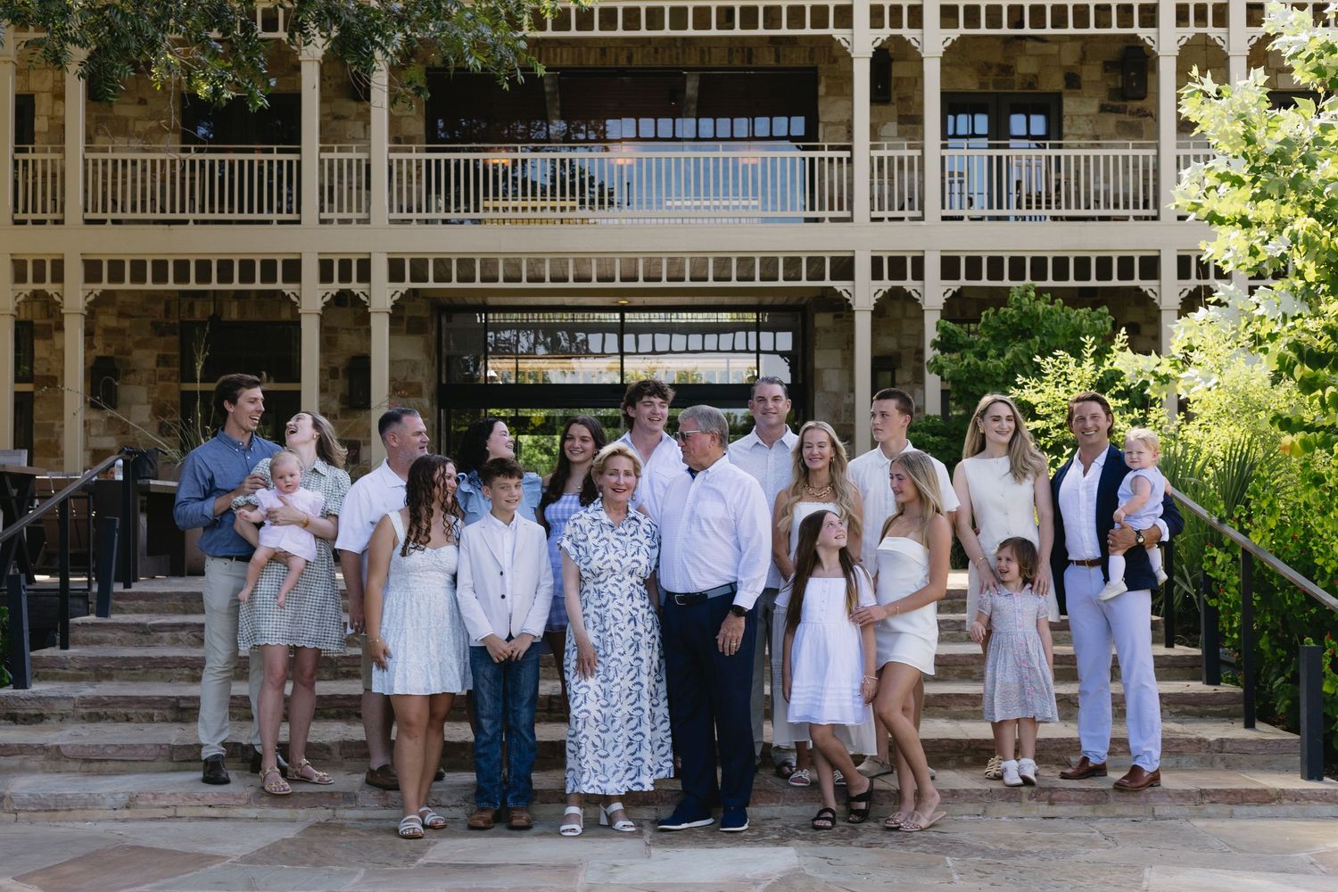 Large family group portrait on stone steps outside an elegant two-story building, dressed in white and blue outfits.