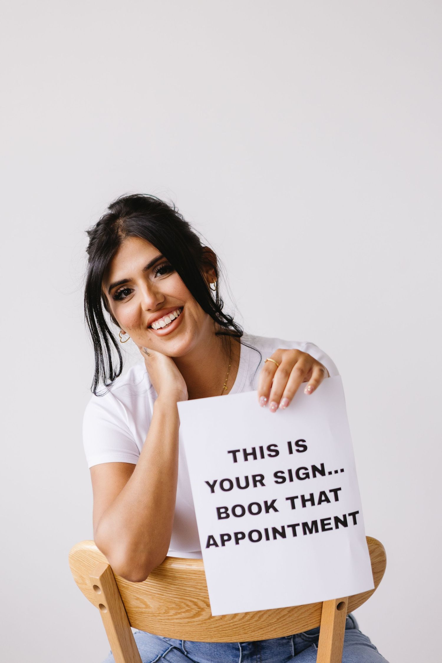 Smiling woman sitting on chair holding sign reading This Is Your Sign Book That Appointment against white background.