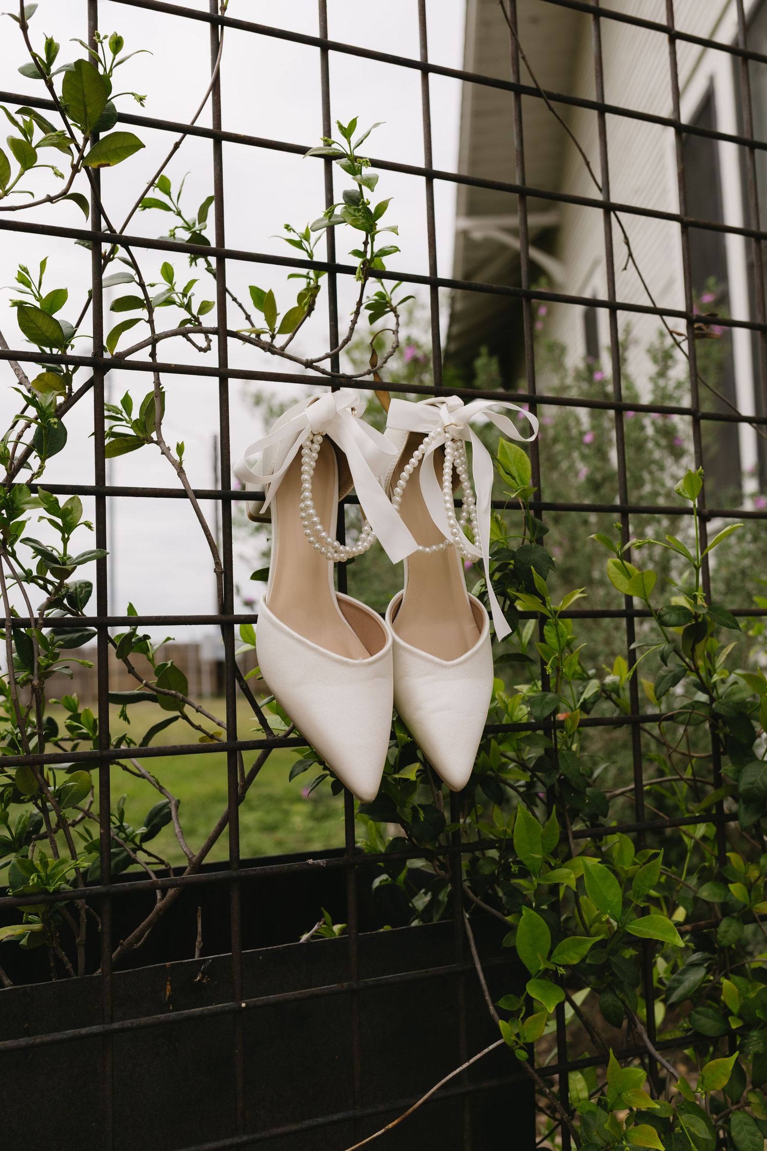 White bridal heels with pearl ankle straps hanging on a black metal grid fence surrounded by green foliage.