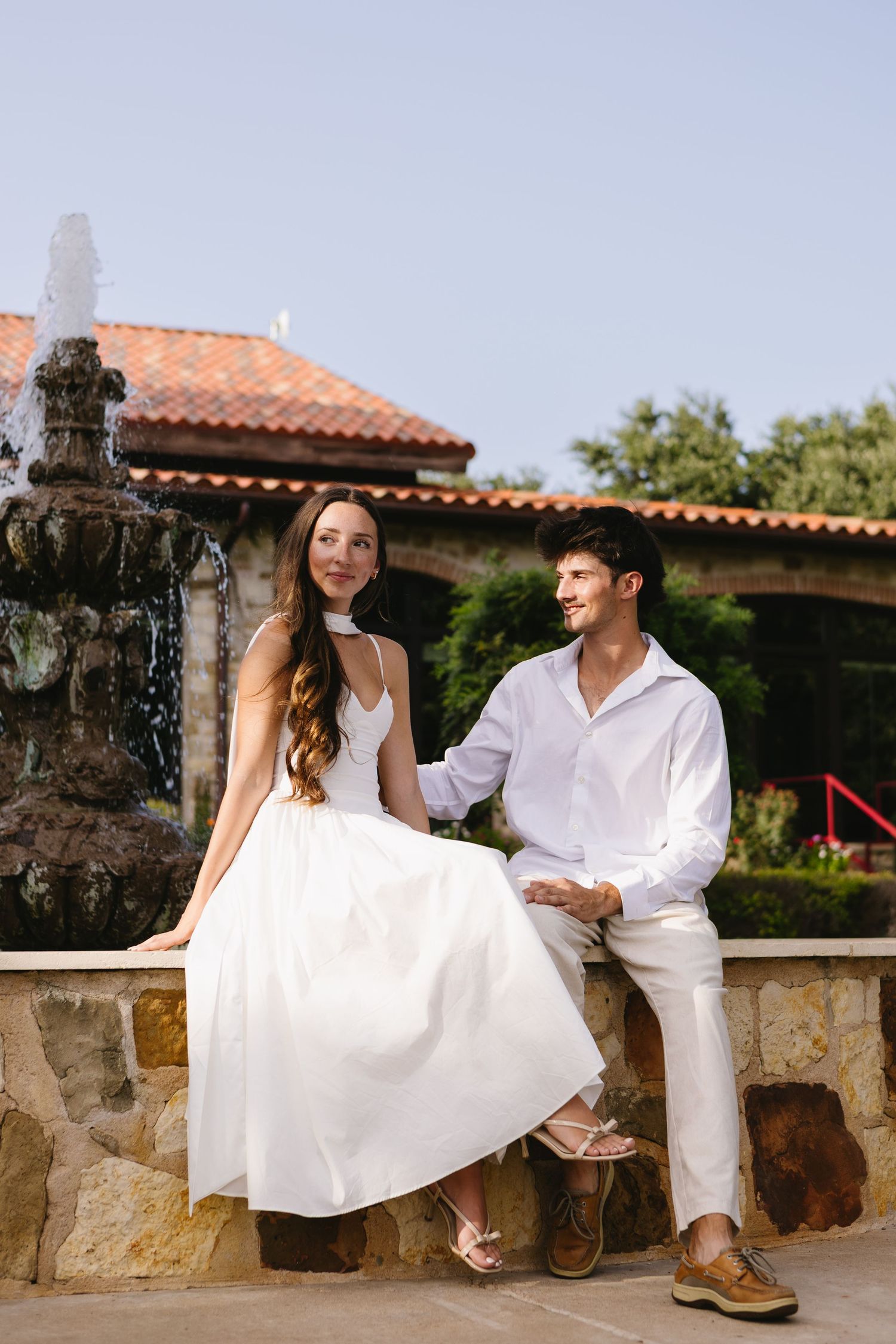 Couple in white attire sitting by a stone fountain outside a Mediterranean-style building on a sunny day.