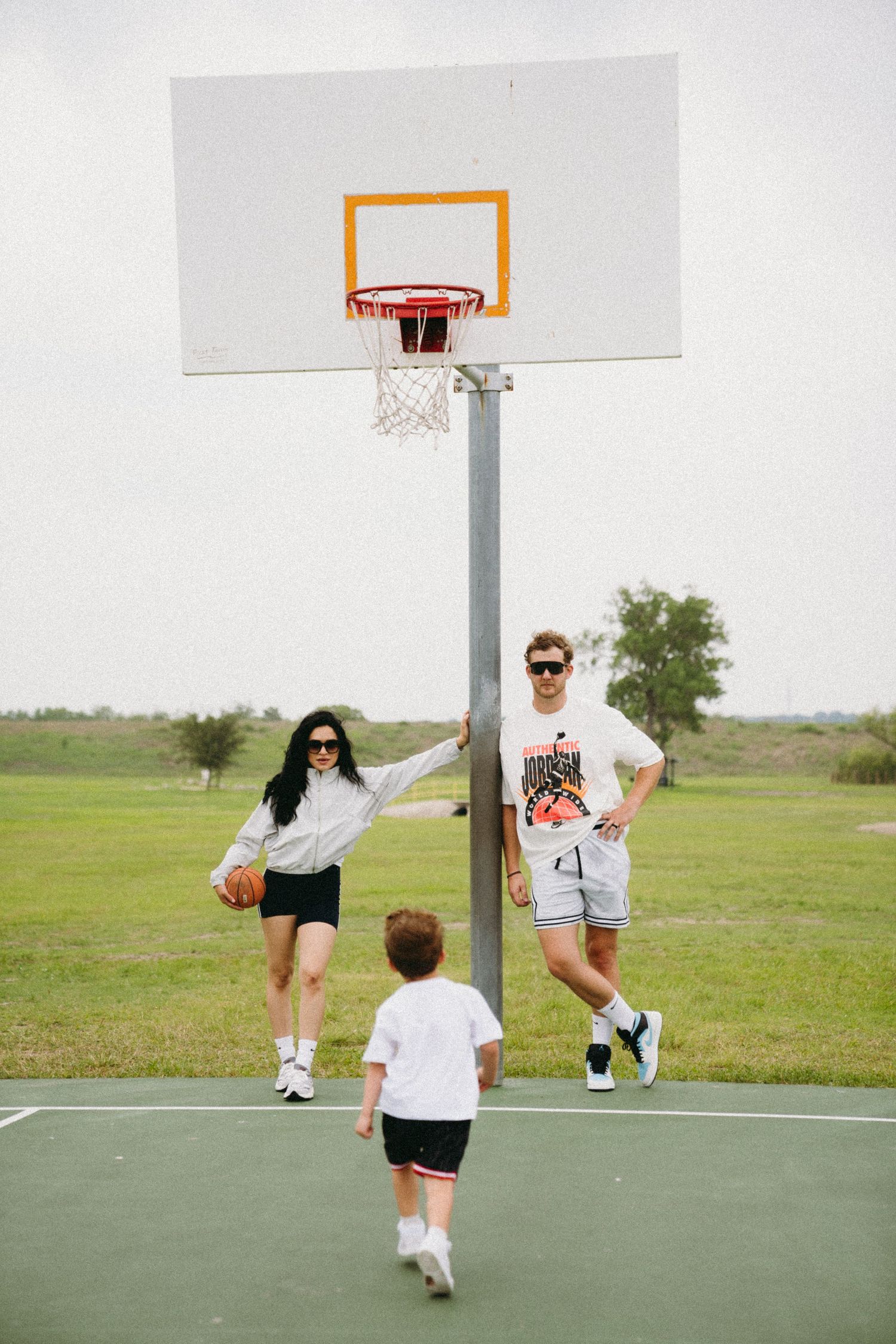 Family playing basketball outdoors on a court, with a woman holding a ball near the hoop and a child running toward them.