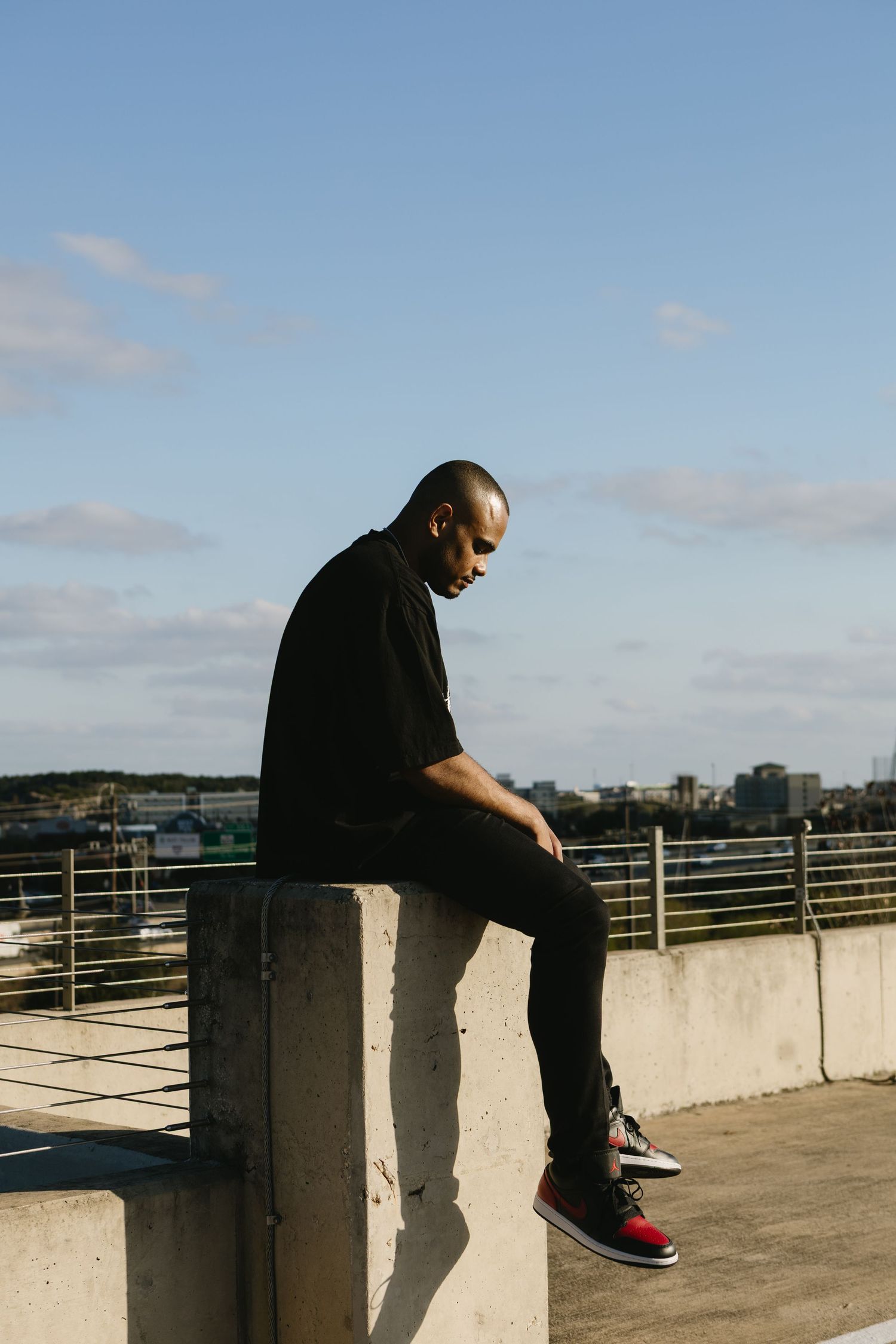 Man in black outfit sitting on concrete wall on rooftop with city skyline and blue sky.