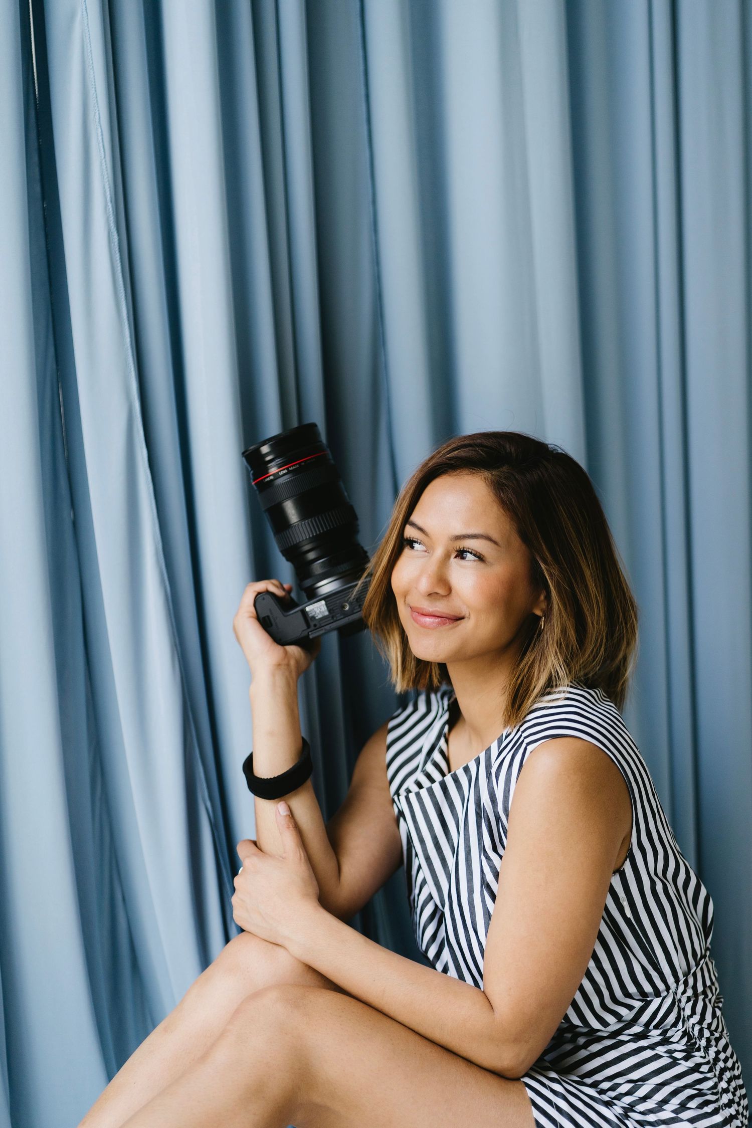 Woman in striped dress holding professional camera against blue curtain backdrop, looking thoughtfully to the side.
