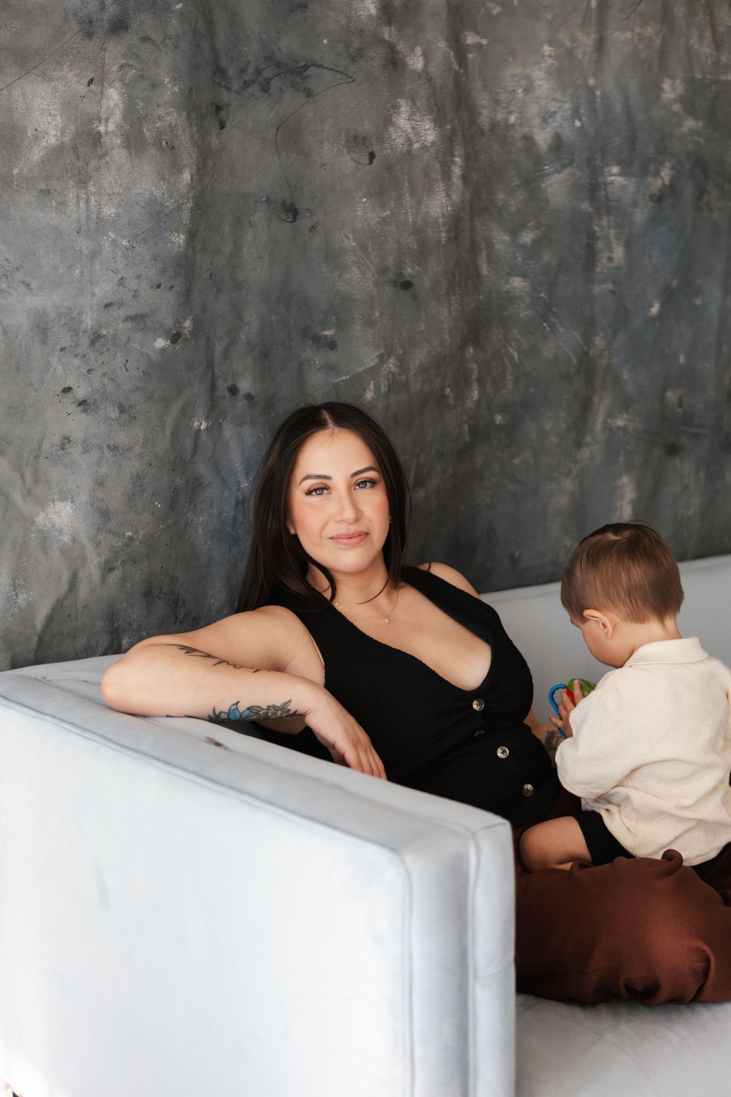 Woman in black dress sitting on white sofa with young child against textured gray wall background.