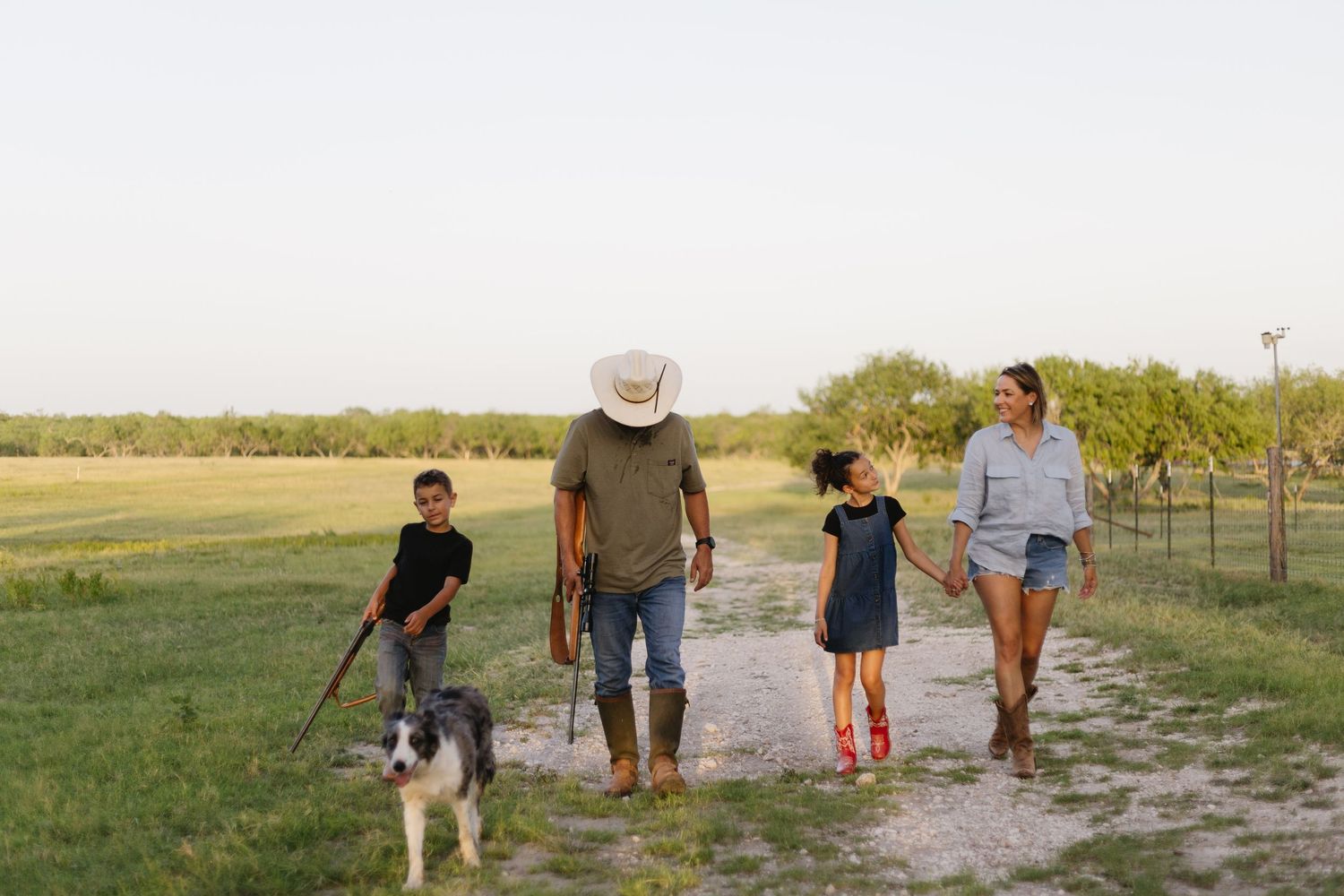 A family walks together with their dog on a dirt path through a rural farm setting at sunset.