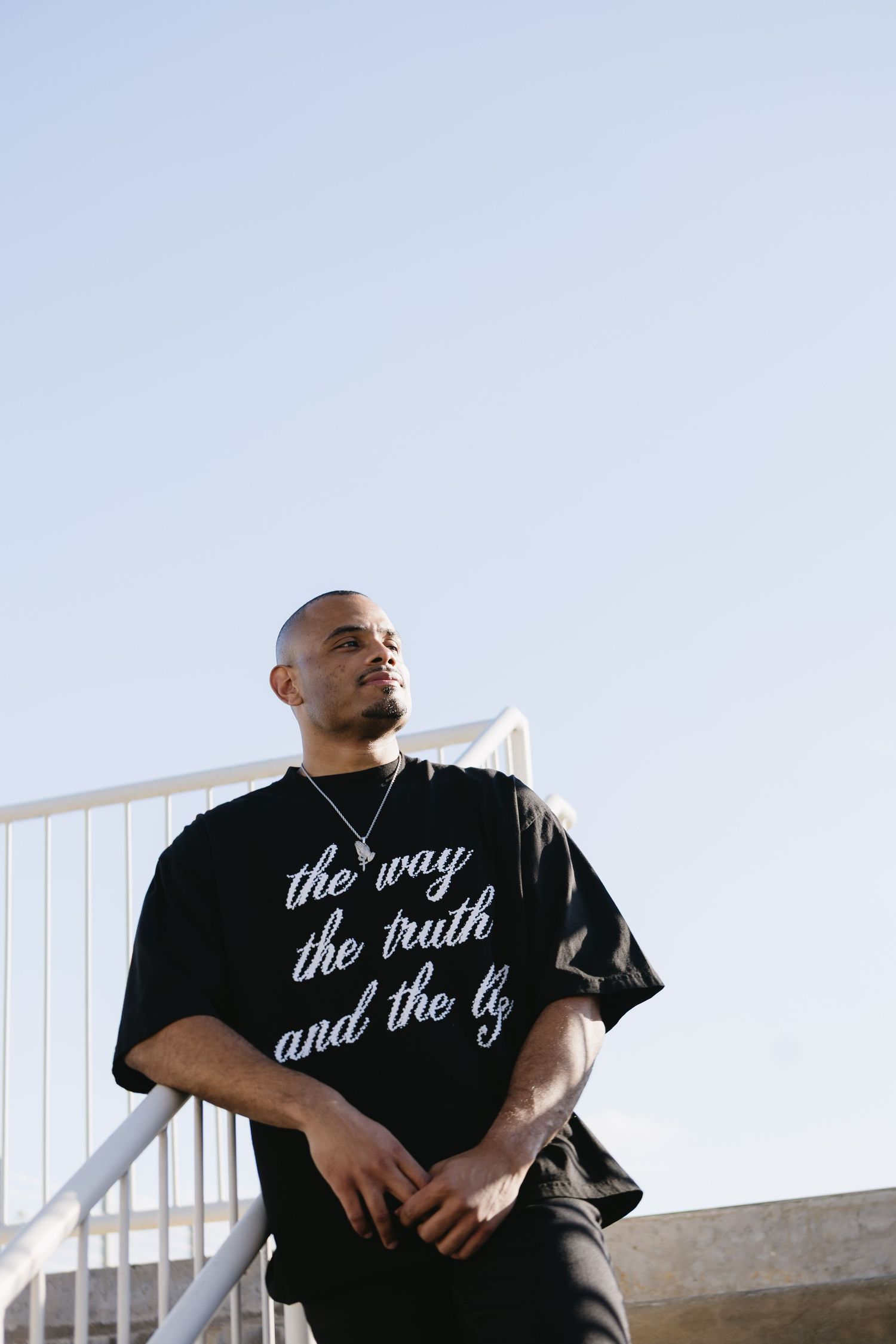 Man in black graphic tee reading 'the way the truth and the life' leaning on railing outdoors.