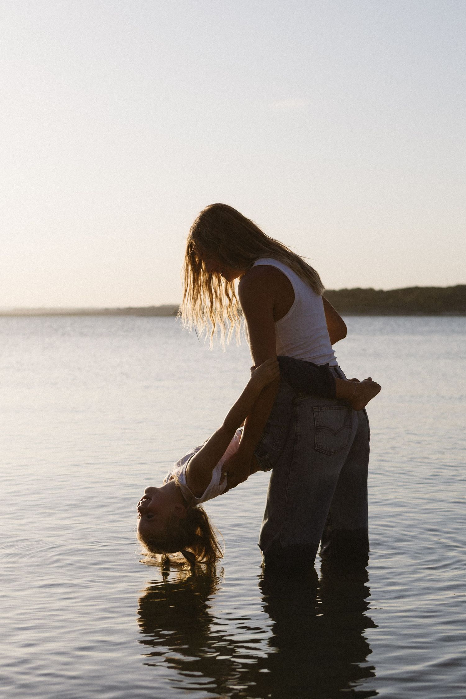 Two women playing in shallow water at sunset, one lifting the other upside down near a calm lake.