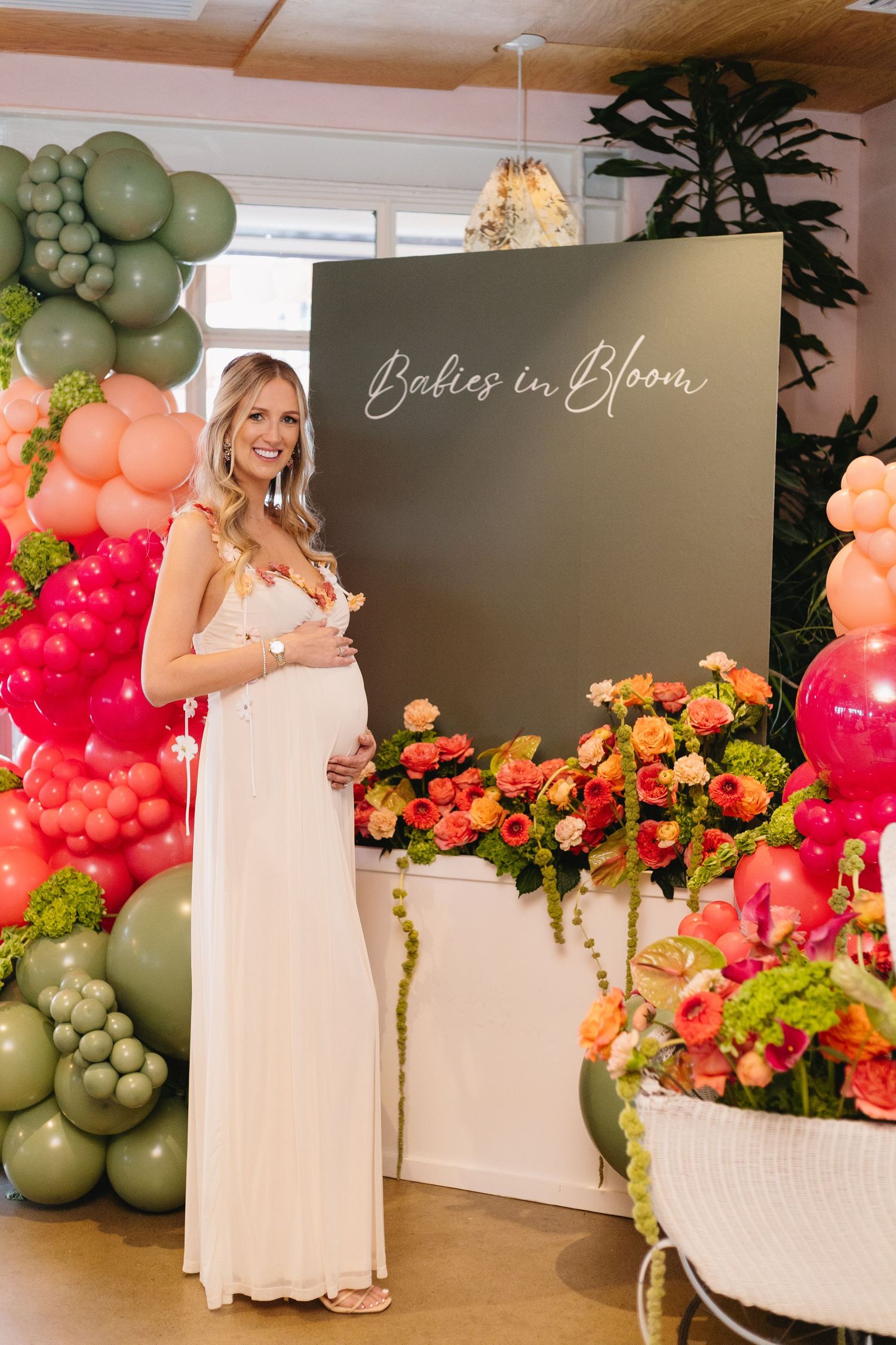 Pregnant woman in white dress at Babies in Bloom baby shower with colorful balloon arch and floral arrangements.