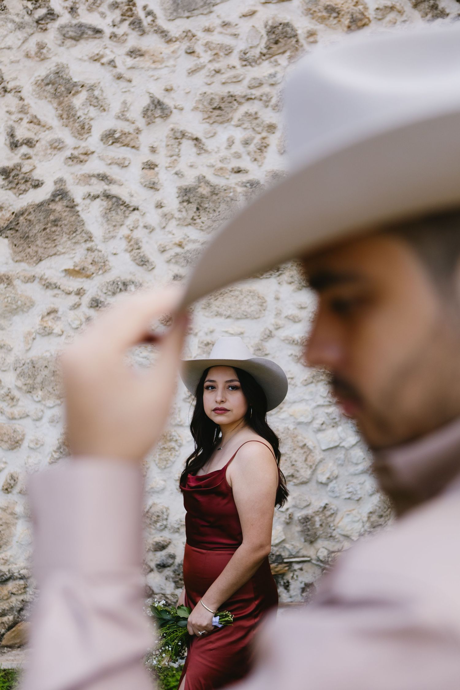 A woman in a burgundy dress and wide-brim hat stands against a stone wall while a man in a cowboy hat poses in the foreground.