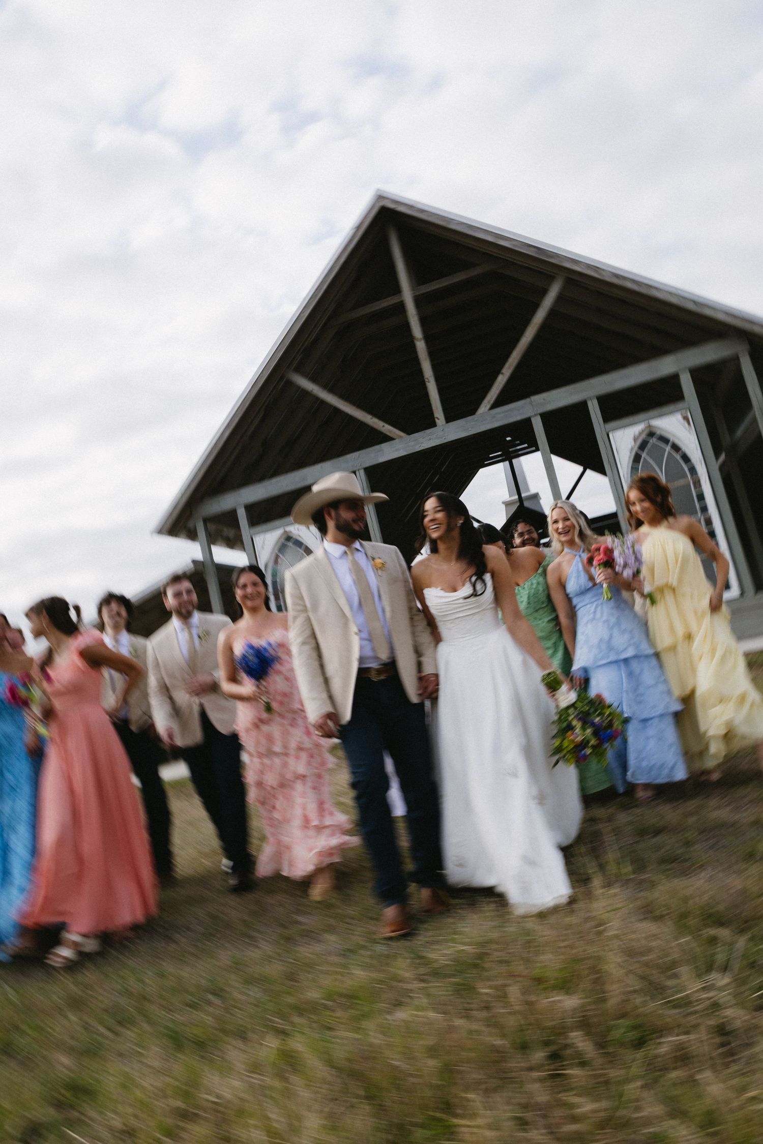 Wedding party walks outdoors near rustic chapel, bride in white gown, groom in tan suit, colorful bridesmaid dresses.