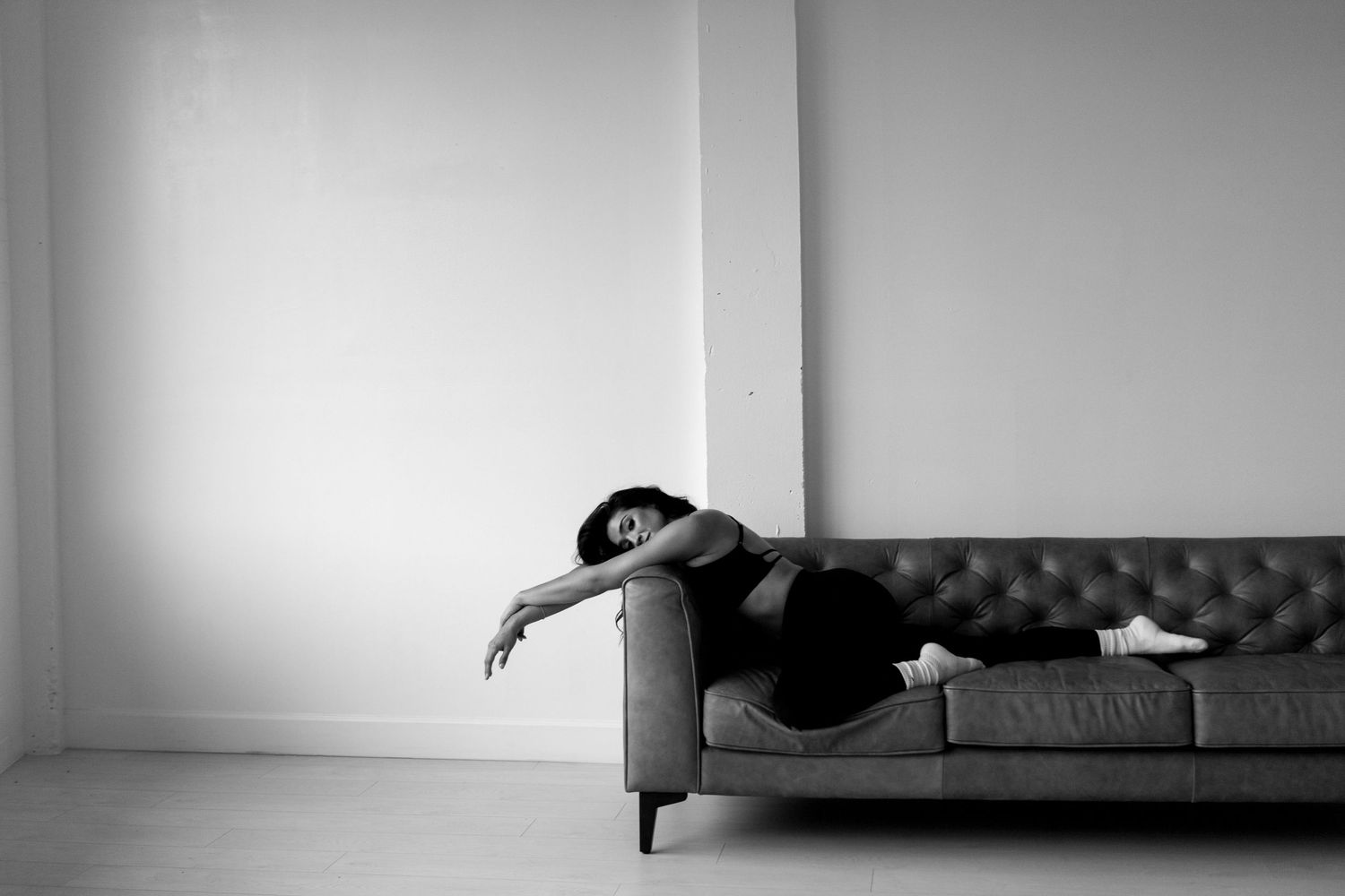 Black and white photo of a woman lying exhausted on a modern tufted sofa in a minimalist room.