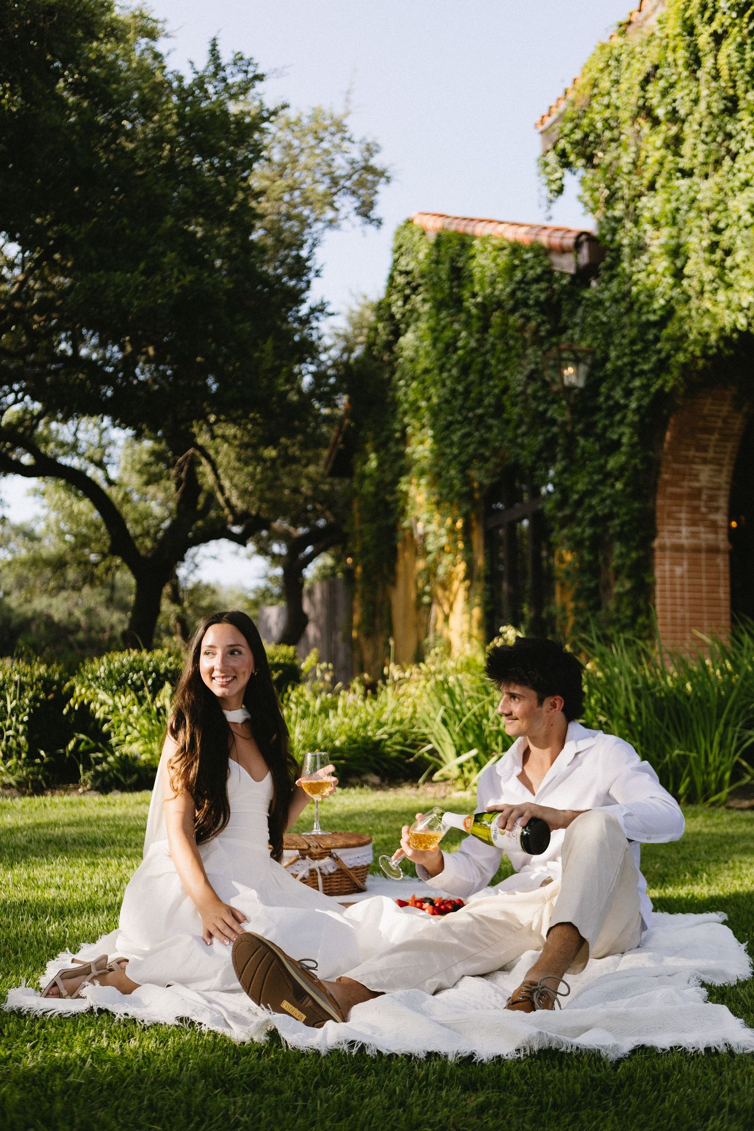 Couple enjoying romantic picnic on lawn with wine and fruit near ivy-covered brick estate.