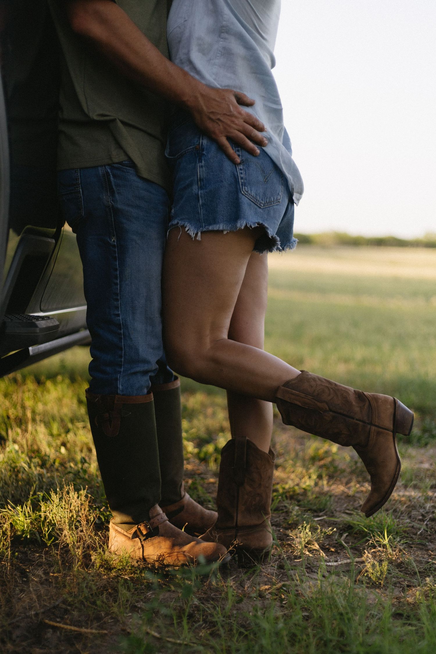 Close-up of a couple in cowboy boots and denim embracing in a sunlit field at sunset.