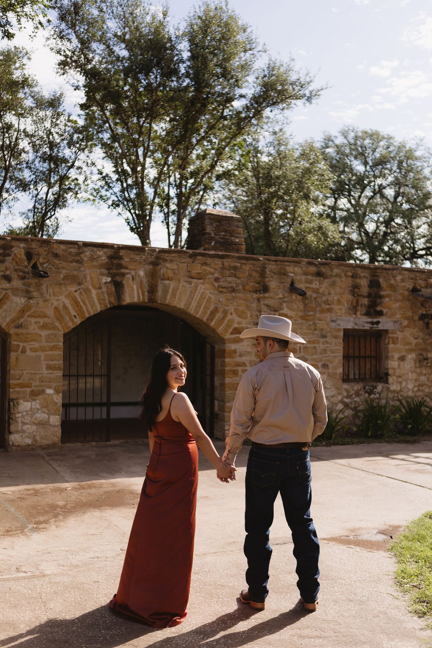 Couple holding hands in front of a rustic stone archway, woman in red dress and man in cowboy hat and jeans.