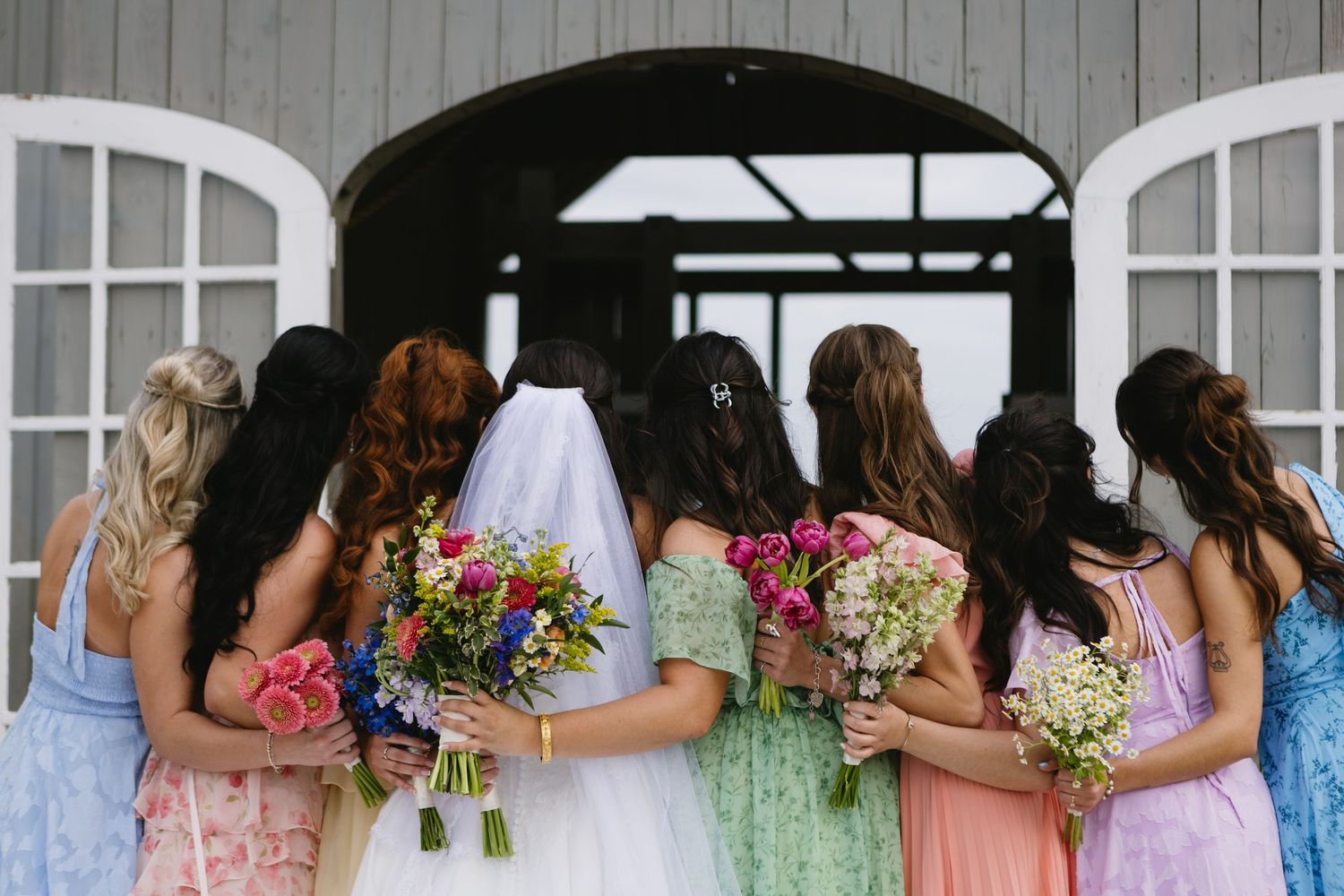 Bride and bridesmaids in colorful dresses holding bouquets, backs to camera, standing in front of barn doors.