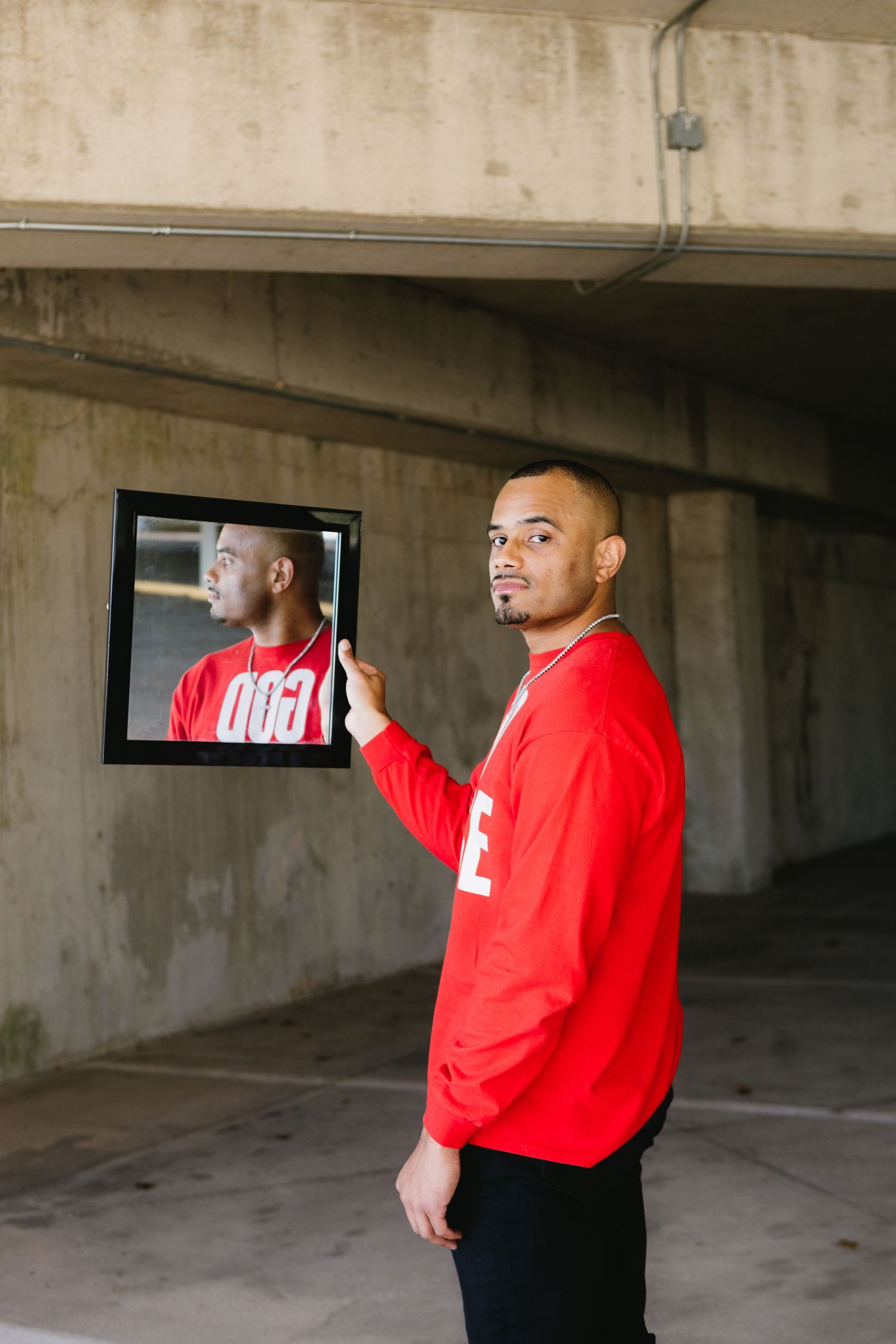 Young man in red GOD CHOSE ME sweatshirt holding framed self-portrait in urban parking structure.