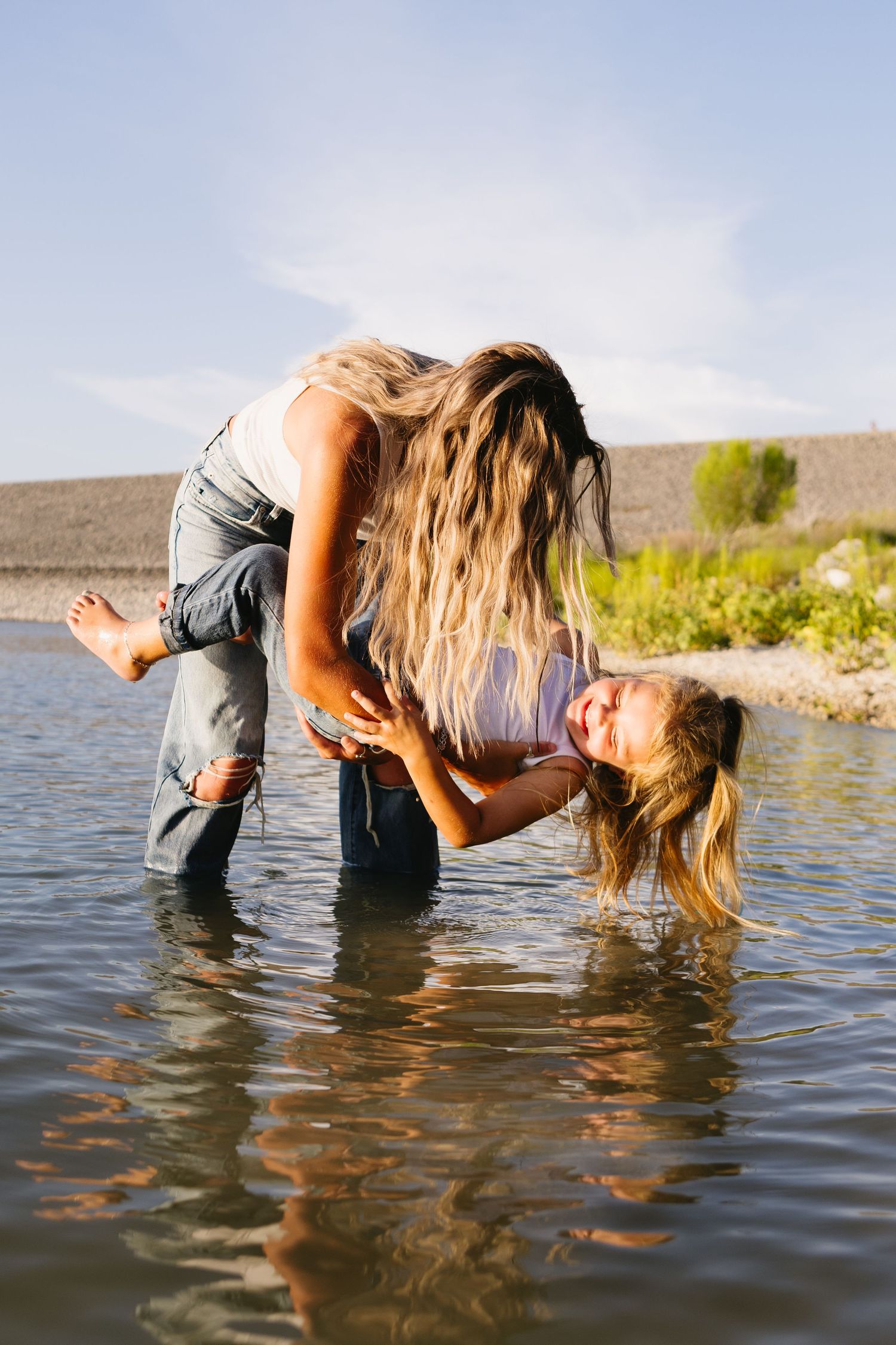 Woman with long hair playfully lifting a laughing child in shallow lake water on a sunny day.