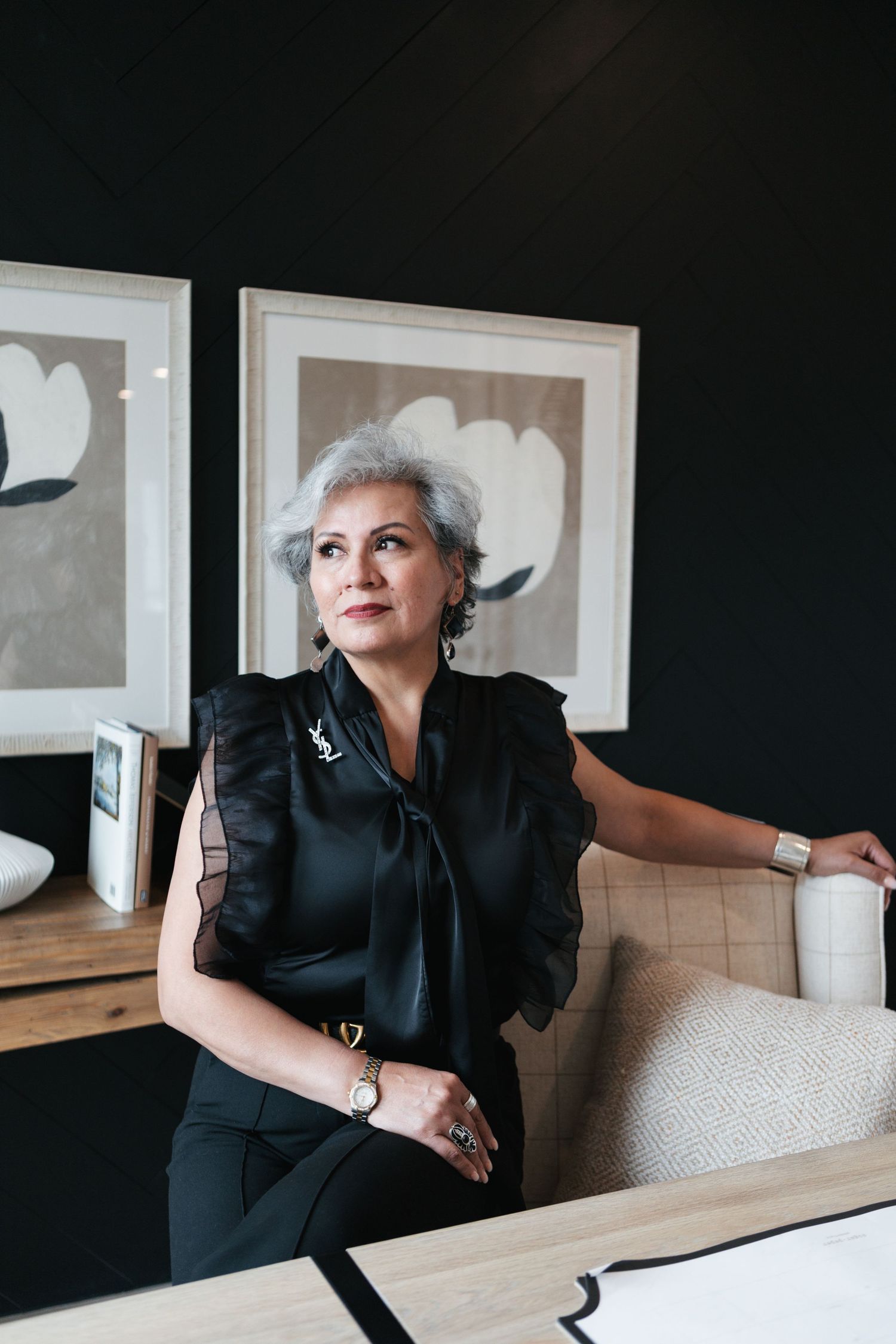 A person in a black outfit sits at a wooden table in a modern interior with white walls and framed artwork.