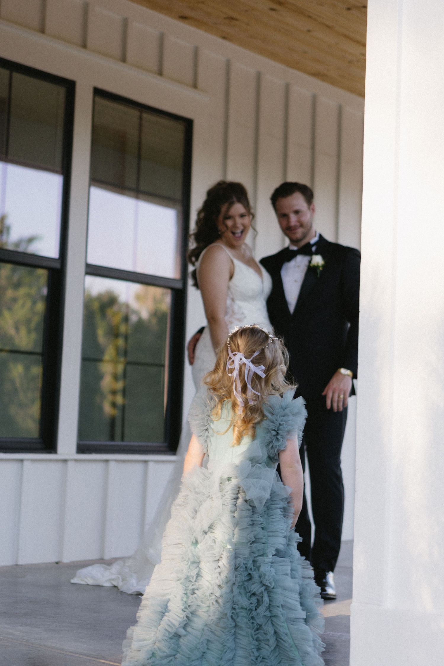 Bride and groom smile on a porch as a flower girl in a ruffled blue dress approaches them at a wedding.