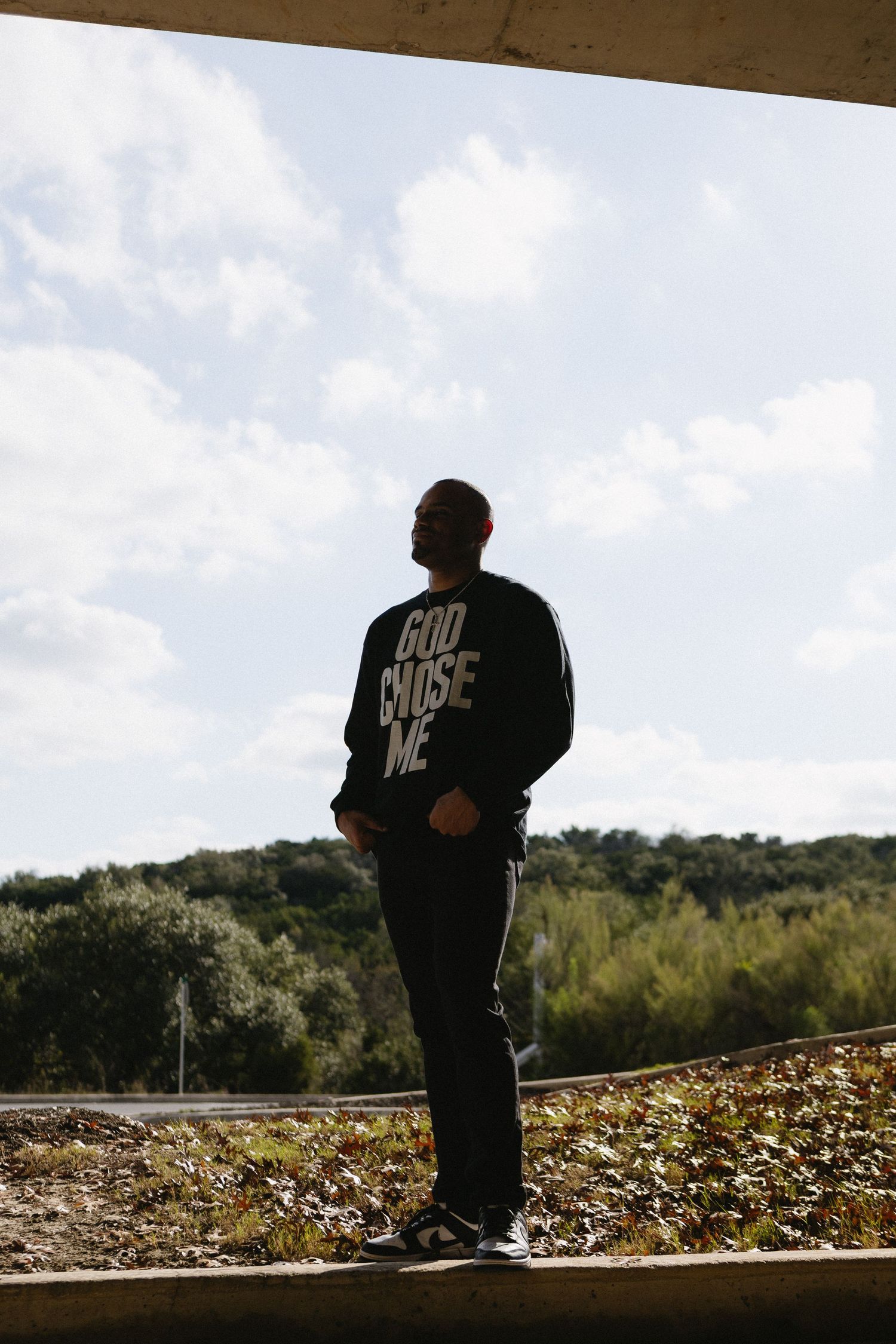 Man in black God Chose Me sweatshirt standing outdoors under overpass with trees and cloudy sky.