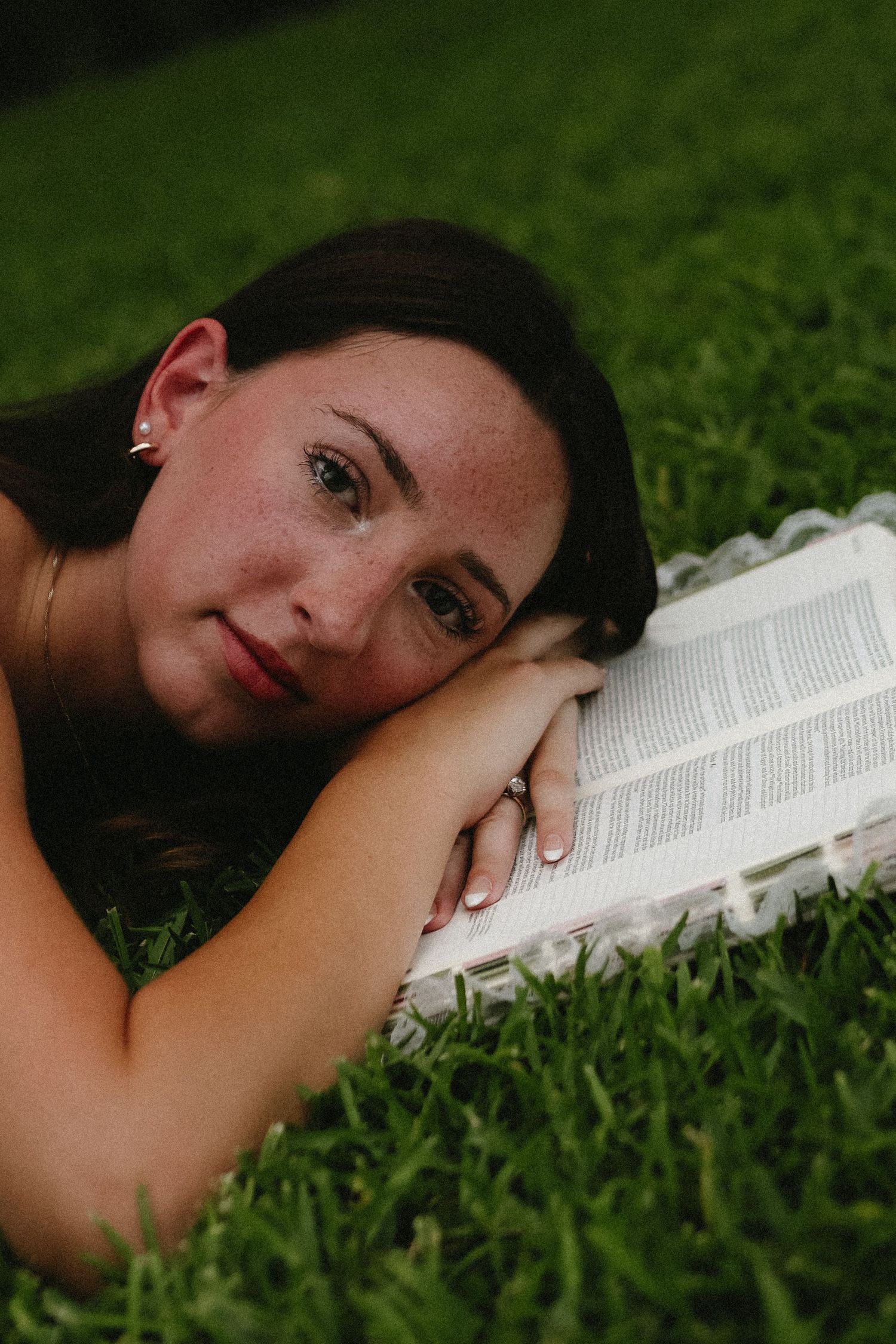 Young woman with freckles resting her head on an open book while lying on green grass outdoors.