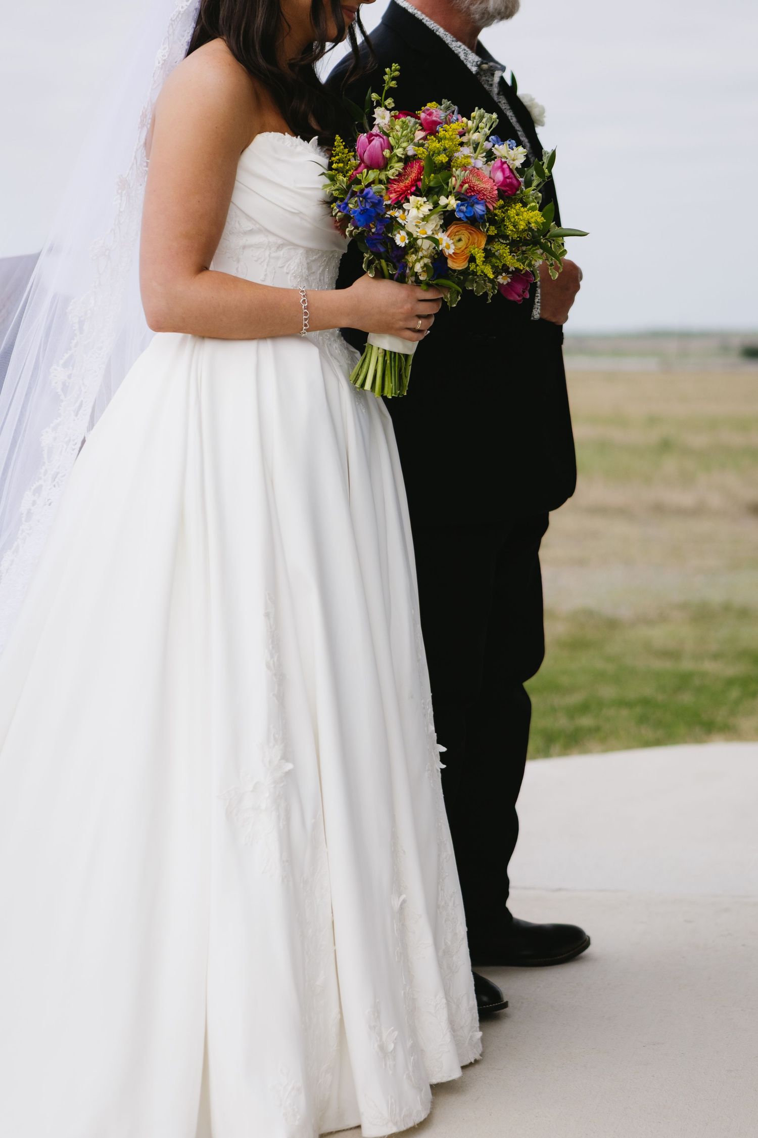 Bride in strapless white gown holds vibrant bouquet beside groom in black suit.