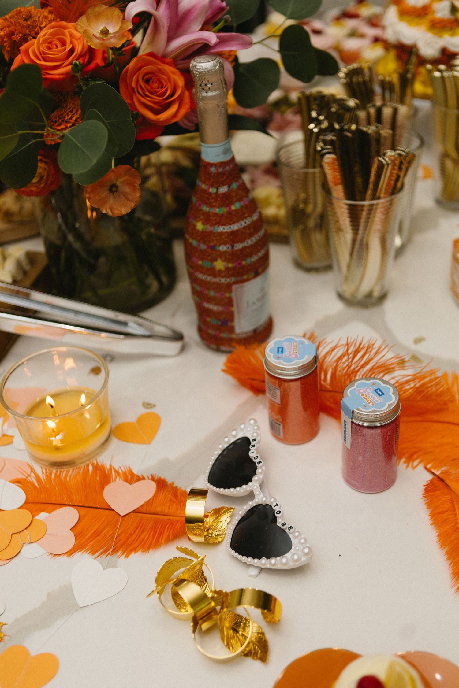 Festive party table with orange roses, champagne, heart sunglasses, feathers, glitter jars, and gold confetti decorations.