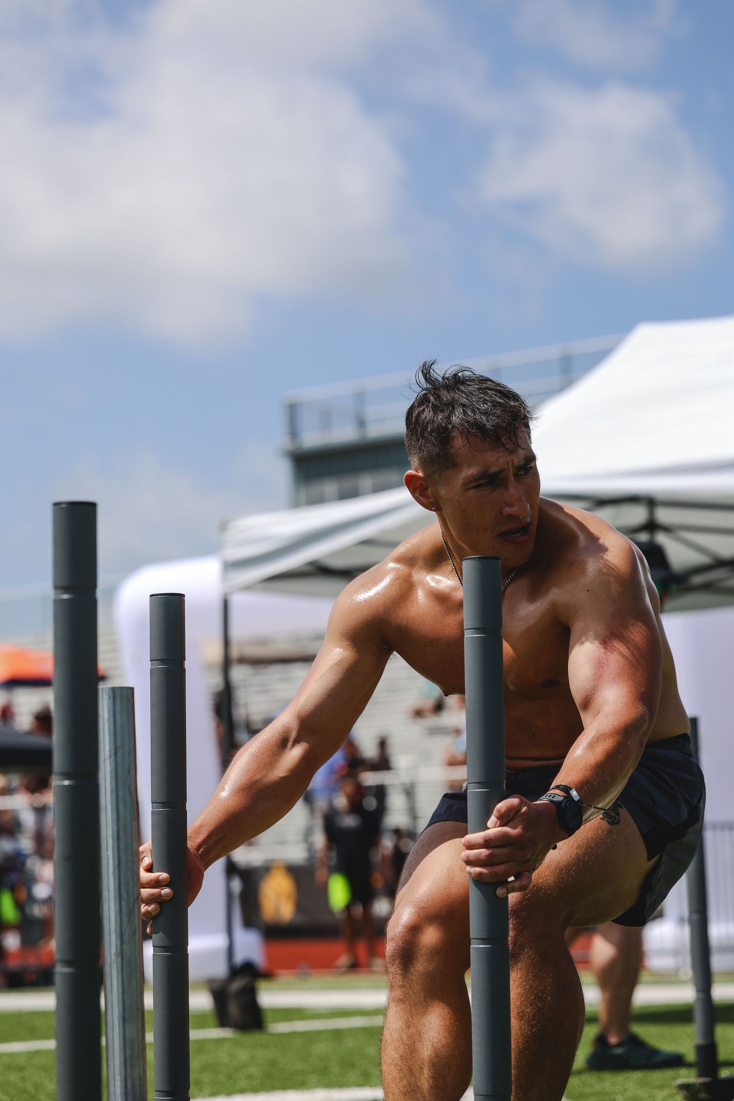 Shirtless muscular man pushes sled on turf at outdoor athletic competition under blue sky.