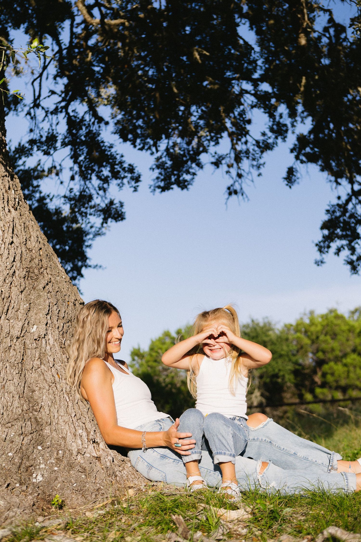 Mother and daughter sitting under a large tree, girl making heart shape with hands, both wearing white tanks and jeans.