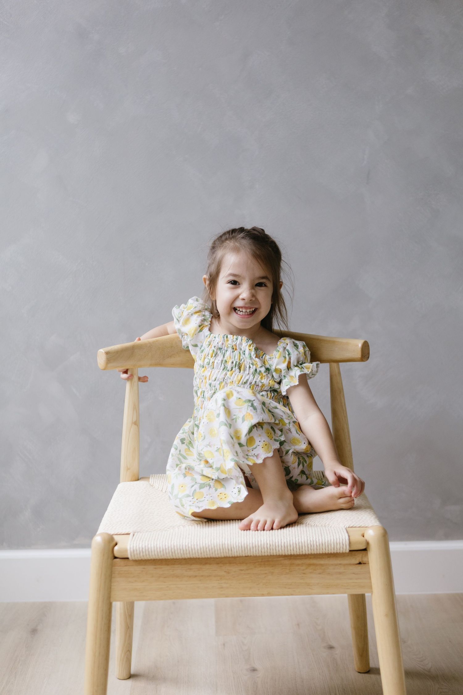A child in a floral dress sits playfully on a wooden chair against a gray wall.