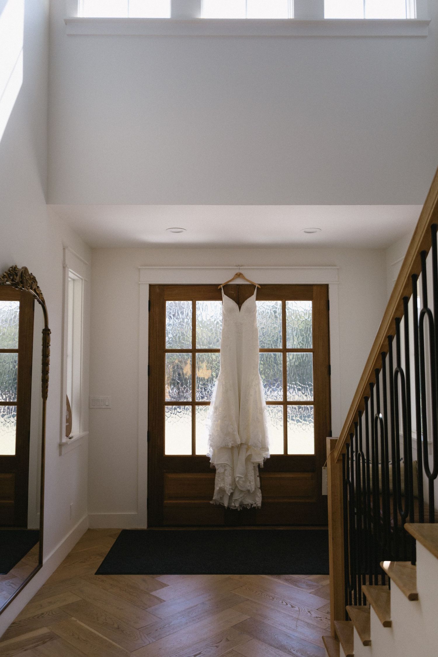 A white wedding dress hanging on a wooden door in an elegant foyer with herringbone floors and a staircase.