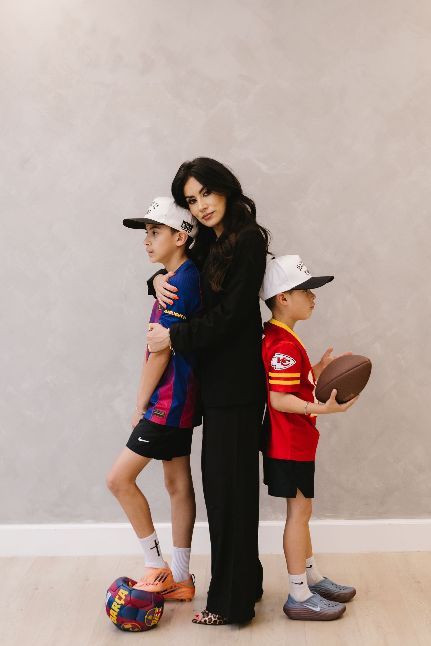 Woman in black outfit poses with two boys in sports jerseys, one holding a football, against a gray wall.