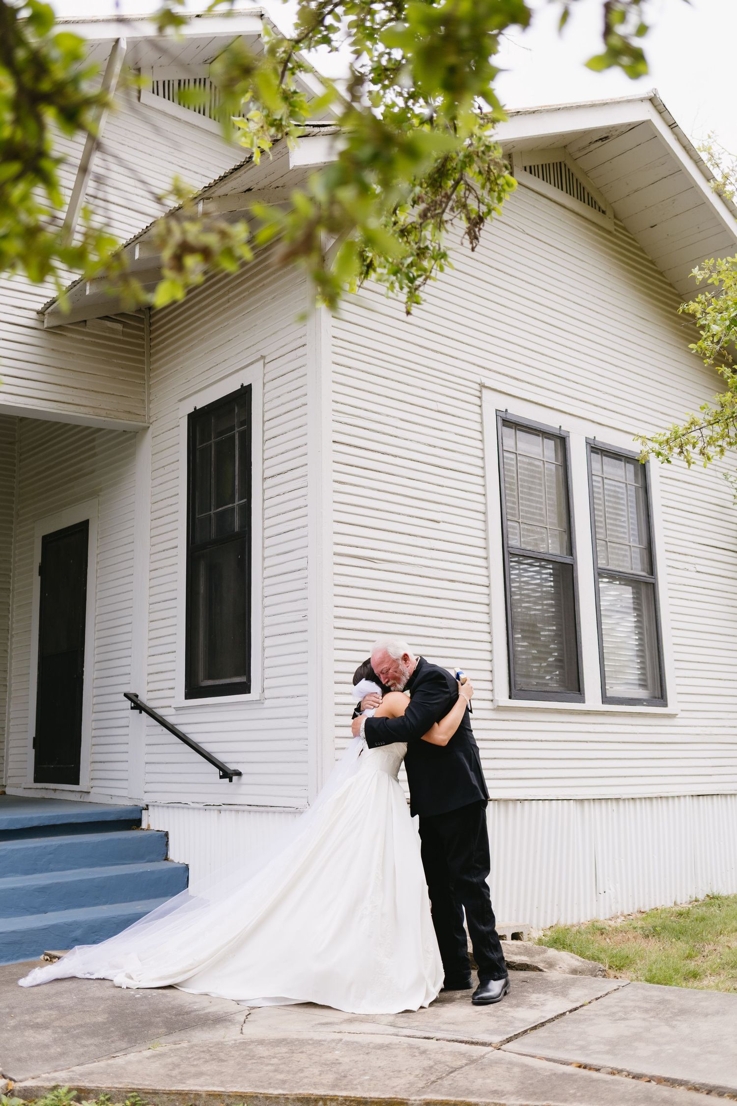 Bride and groom share a romantic embrace outside a white wooden church on their wedding day.