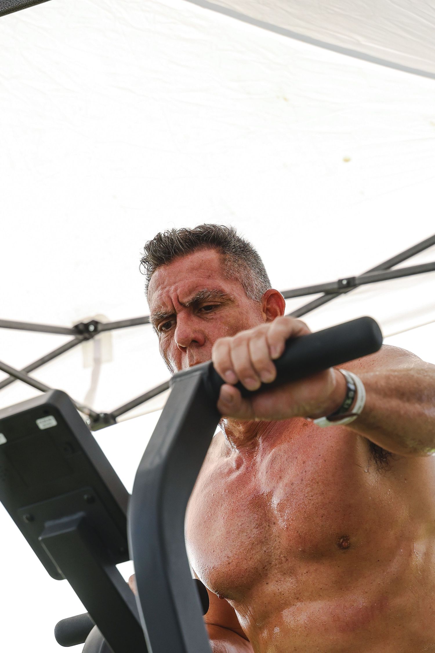 Muscular shirtless man exercising on an elliptical machine outdoors under a canopy, sweating during an intense workout.