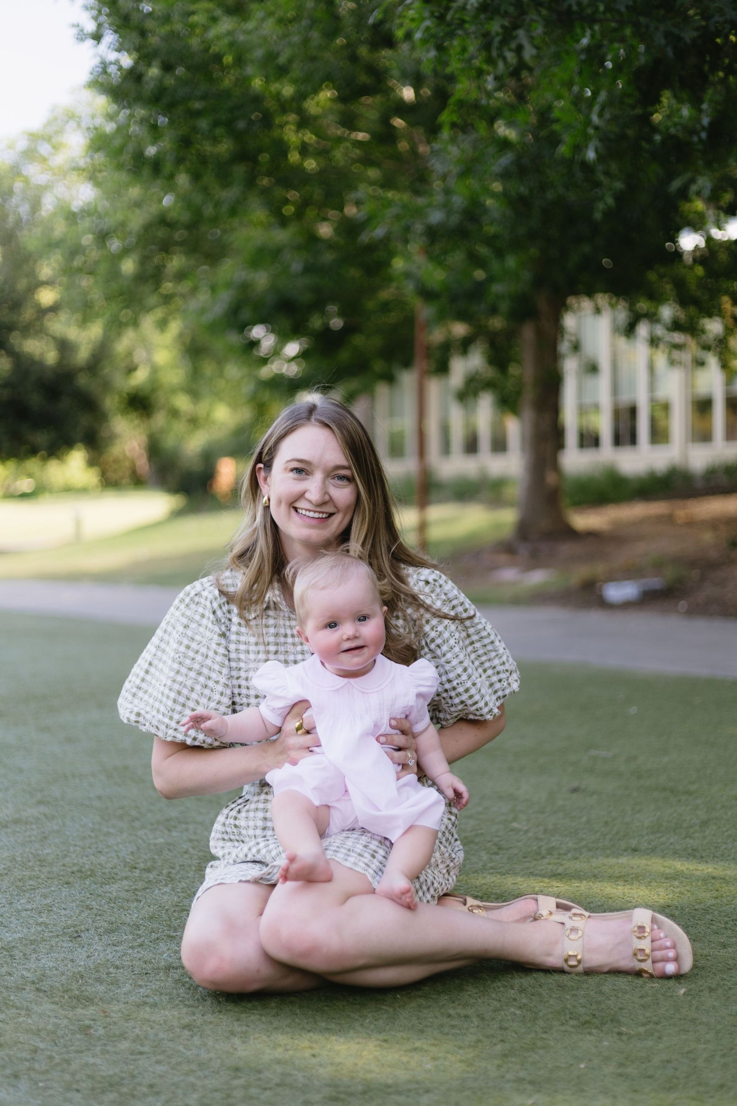Smiling woman sitting cross-legged on green grass holding a baby in a white outfit outdoors.