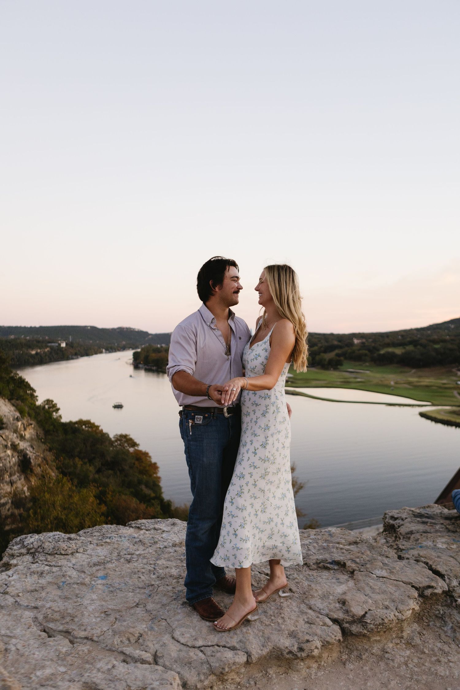 A couple shares a romantic moment on a rocky cliff overlooking a serene lake at sunset.