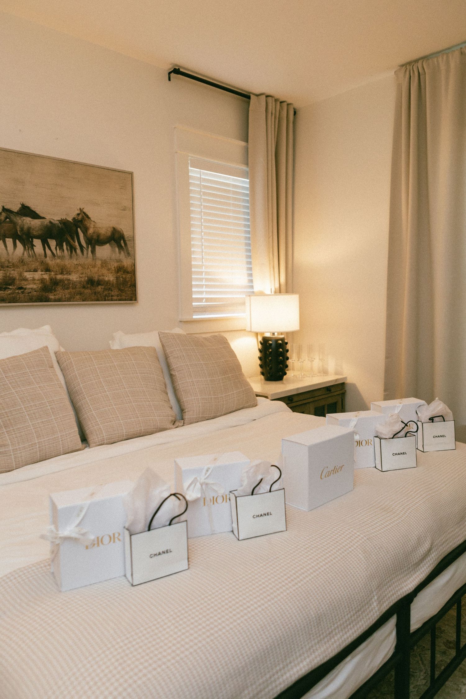 Luxury gift bags from Dior, Chanel, and Cartier arranged on a white bed in an elegant neutral-toned bedroom.