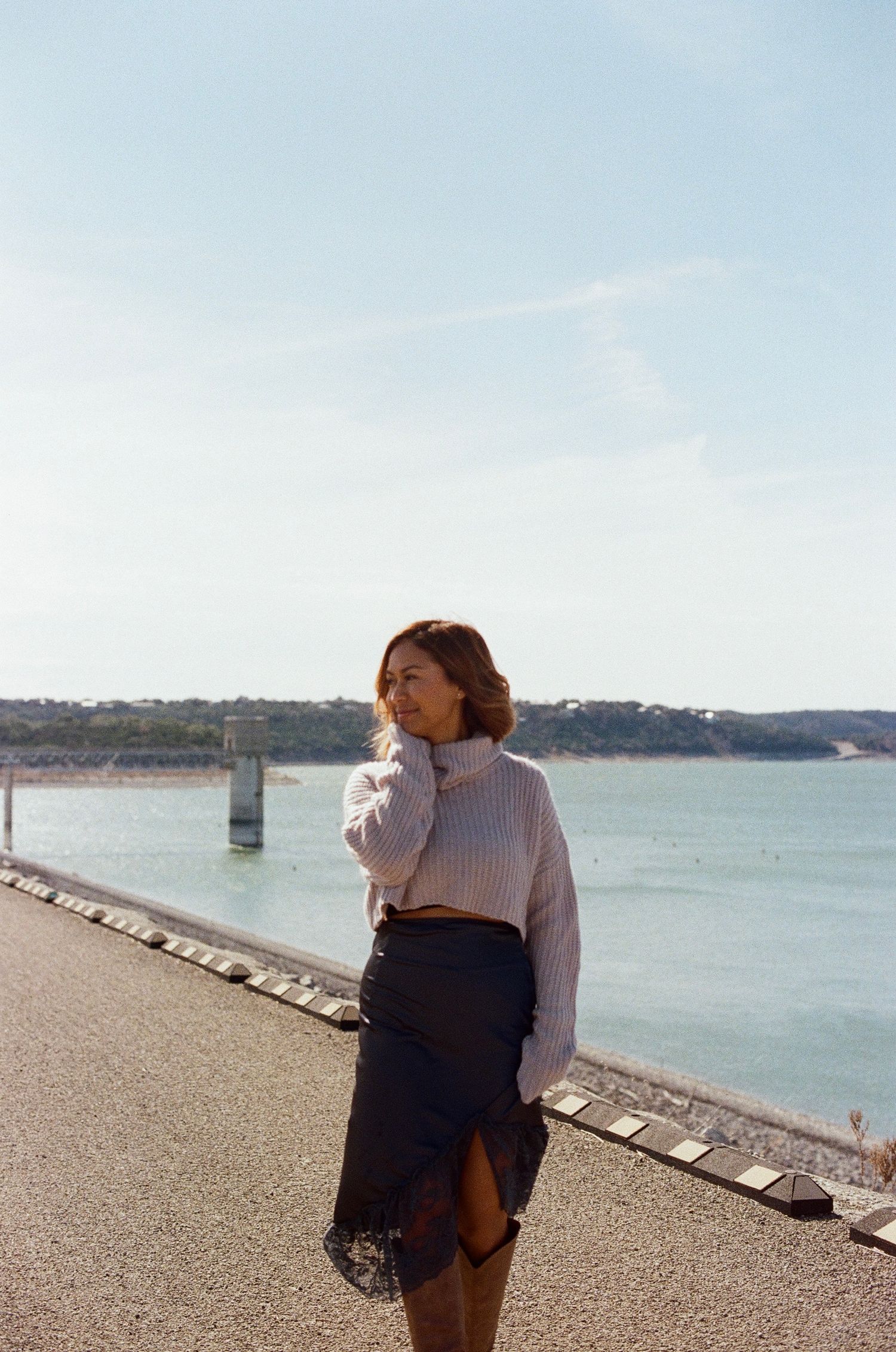 Woman in white sweater and dark skirt stands by waterfront on sunny day, gazing into distance.