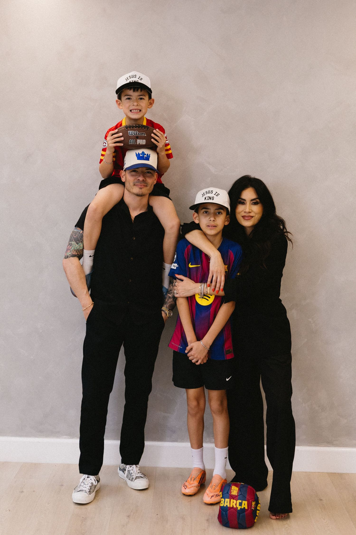 Family of four posing together, man carrying young boy on shoulders, woman hugging older child in FC Barcelona jersey.