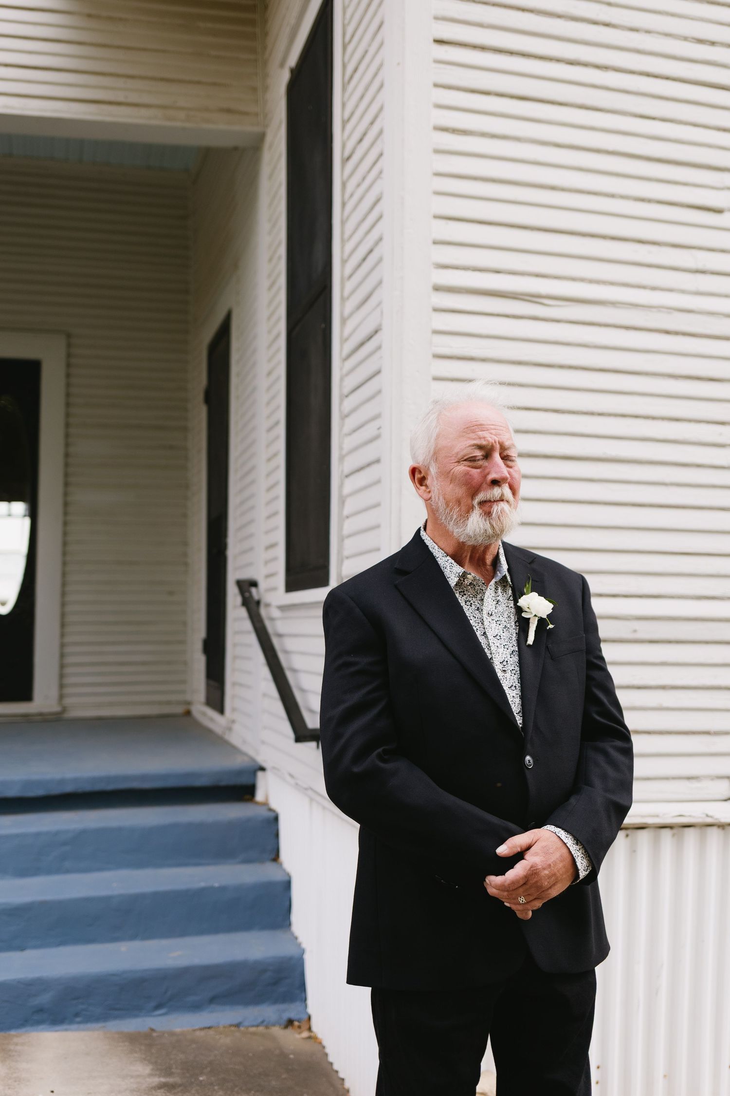 Elderly man in black suit with white boutonniere stands outside vintage building, dressed for a wedding celebration.