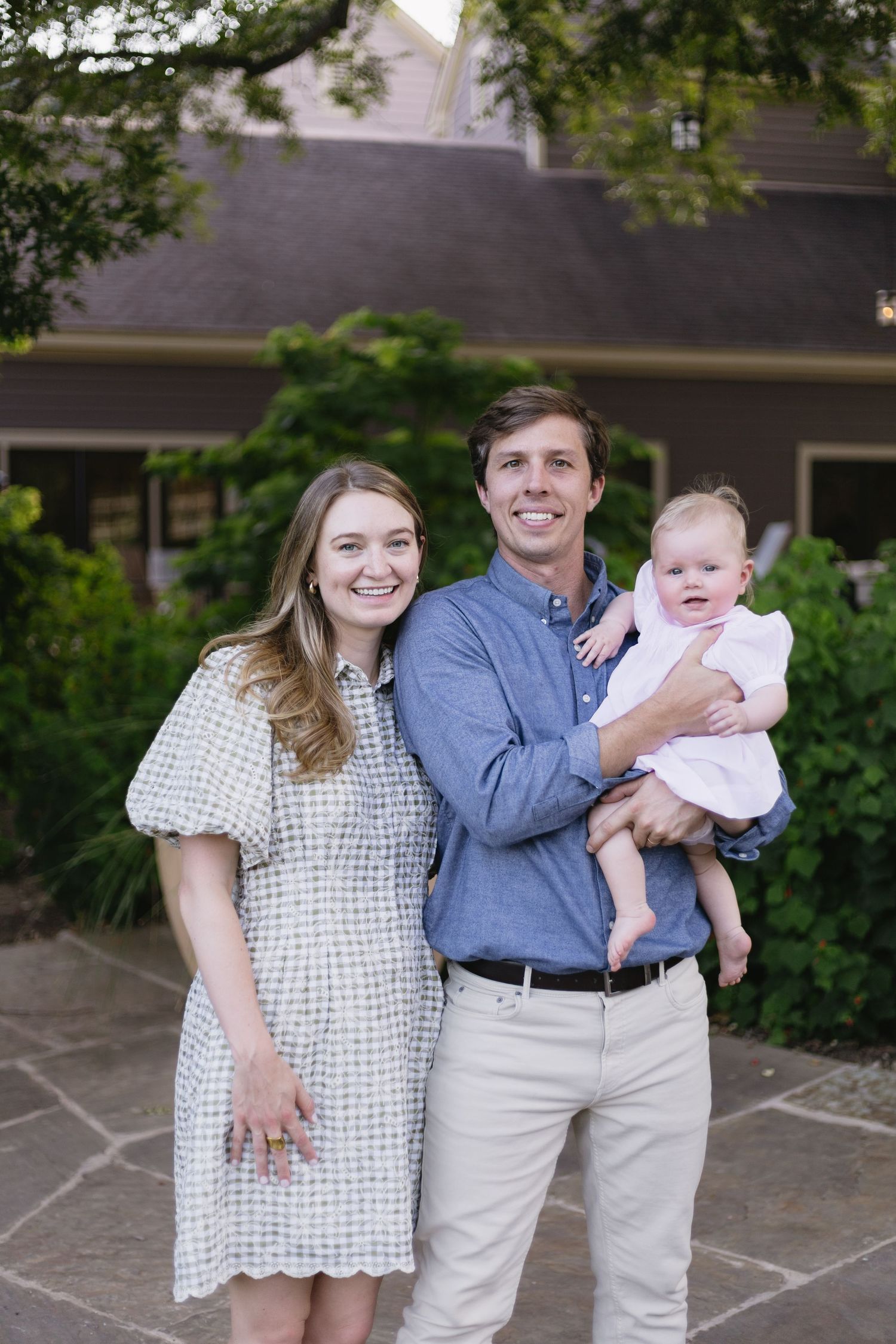 Young family smiling outdoors, man holding baby girl, woman in gingham dress, posing in front of a home.
