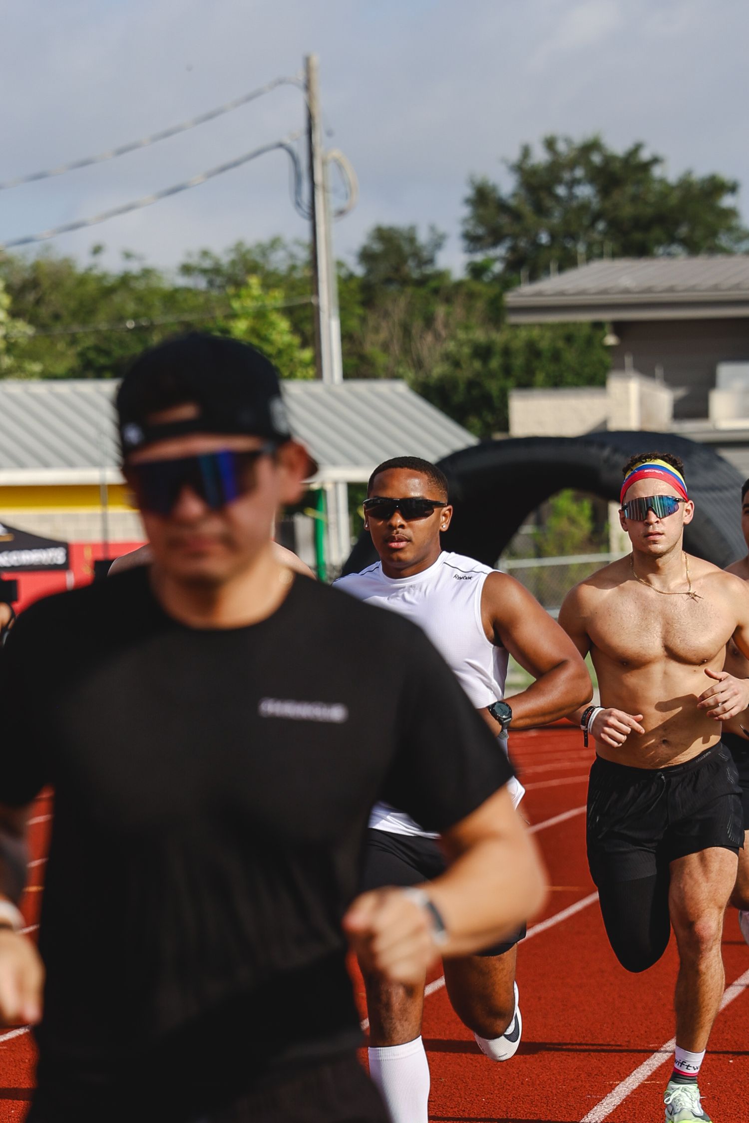 Three athletes running on a red outdoor track, wearing sunglasses and athletic gear on a sunny day.